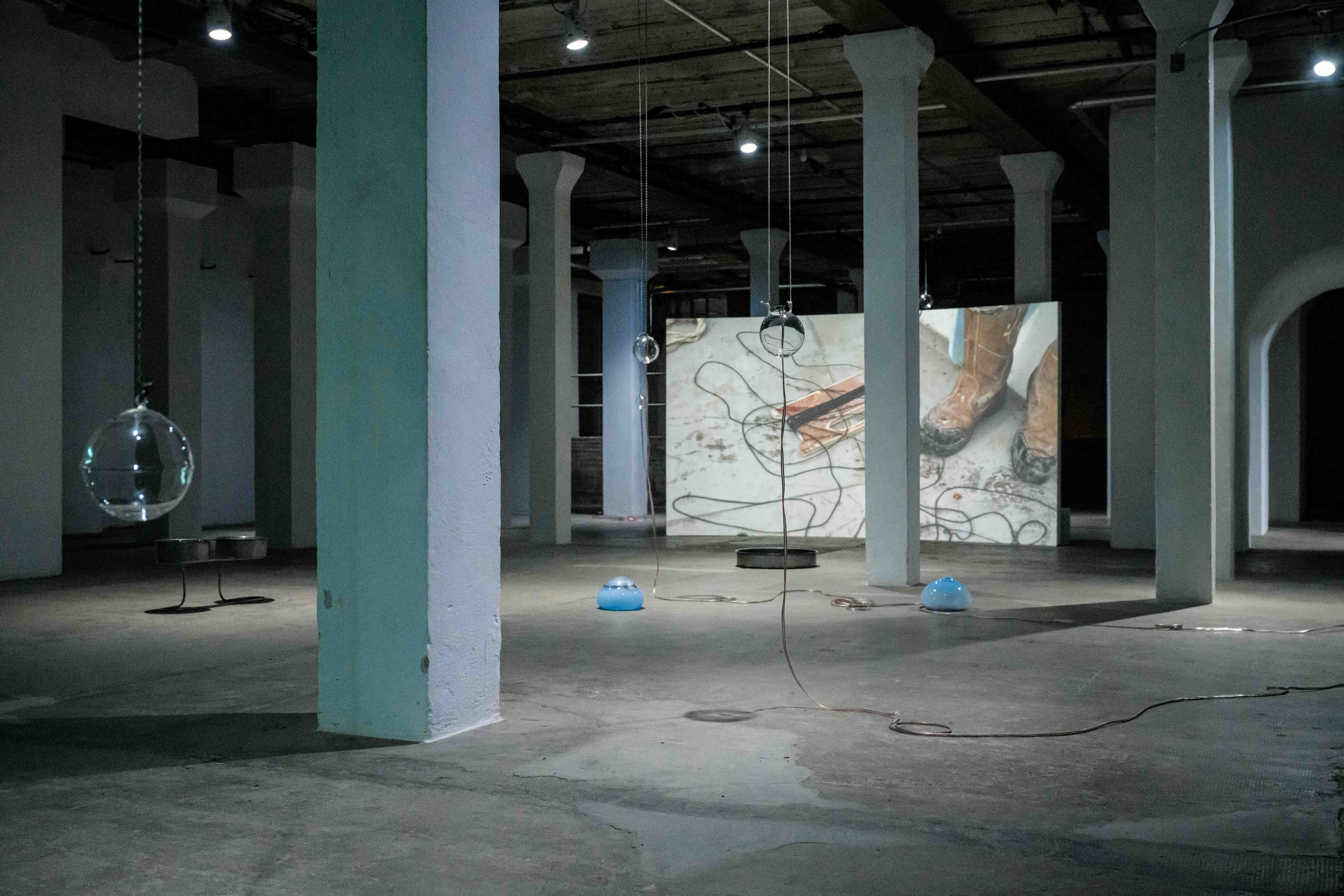 A dimly lit installation by Steph Huang with white pillars displays suspended glass orbs, blue dome objects on the floor, and scattered cables. A large video projection on the far wall shows muddy boots and tangled wires, adding to the industrial atmosphere.