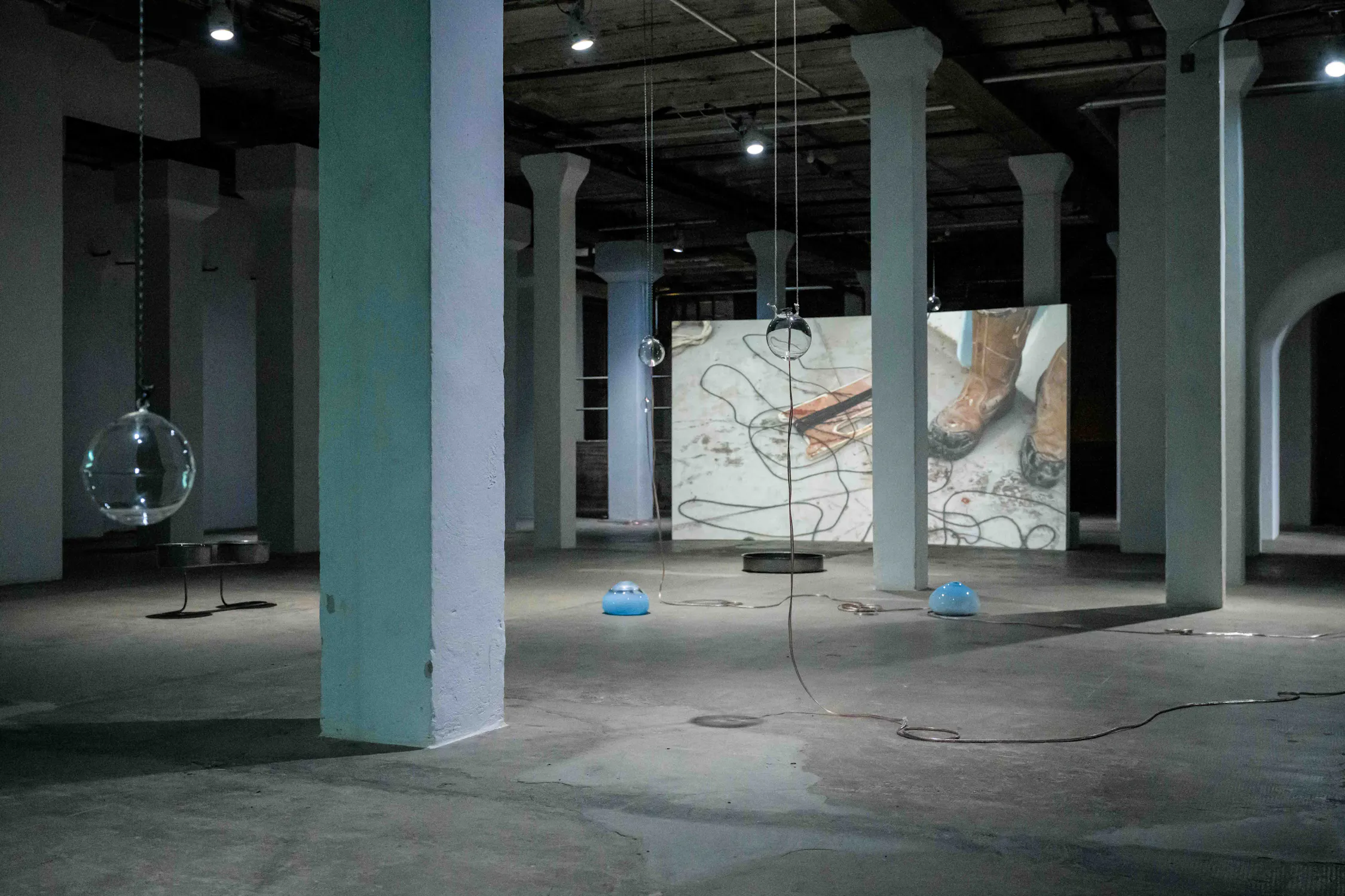 A dimly lit installation by Steph Huang with white pillars displays suspended glass orbs, blue dome objects on the floor, and scattered cables. A large video projection on the far wall shows muddy boots and tangled wires, adding to the industrial atmosphere.