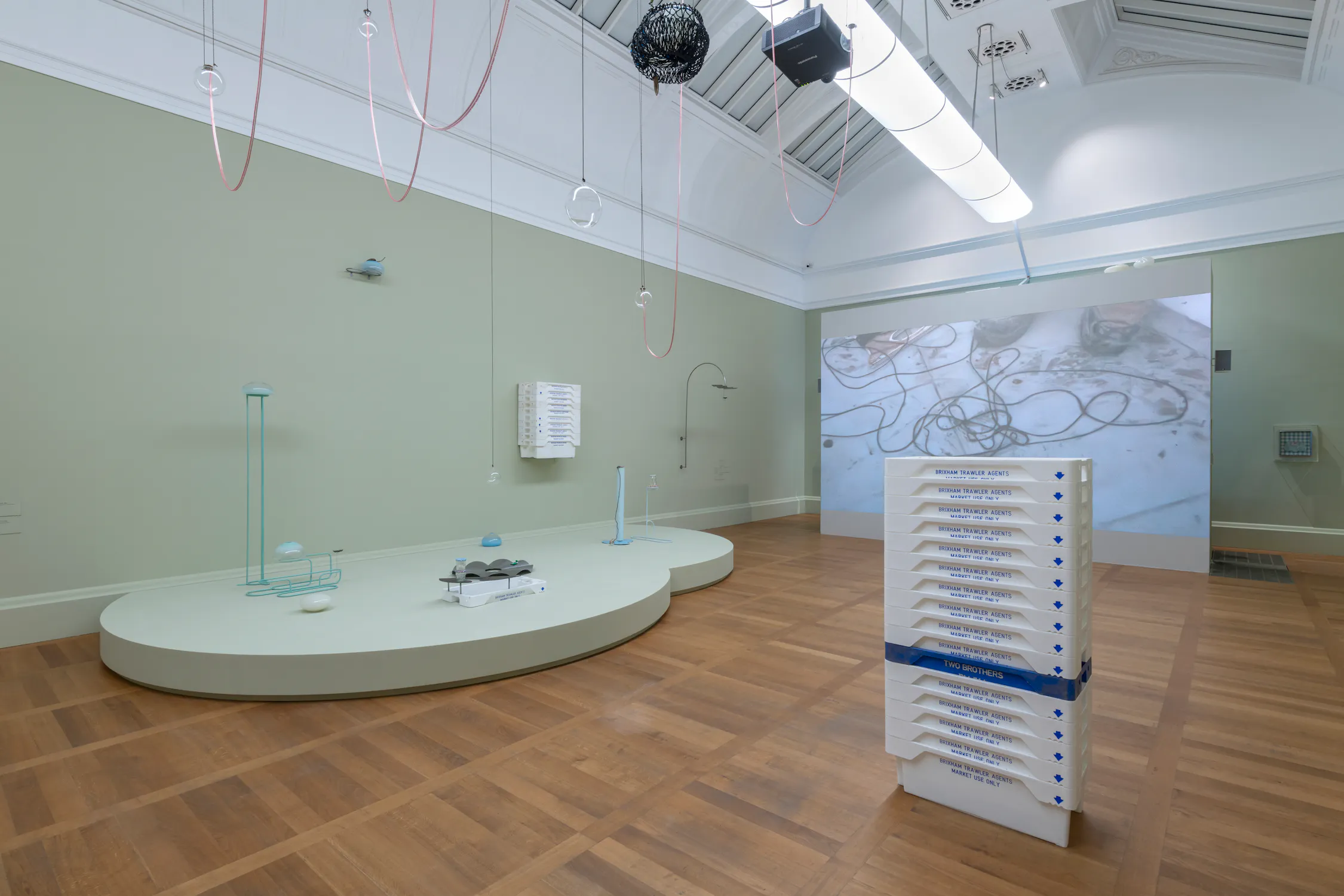 A contemporary installation by Steph Huang in contemporary art gallery with wooden floors displays abstract sculptures and installations, including a pale green platform with minimalist objects, a tall stack of white trays, hanging wires, and a large projection of tangled cables on the back wall.