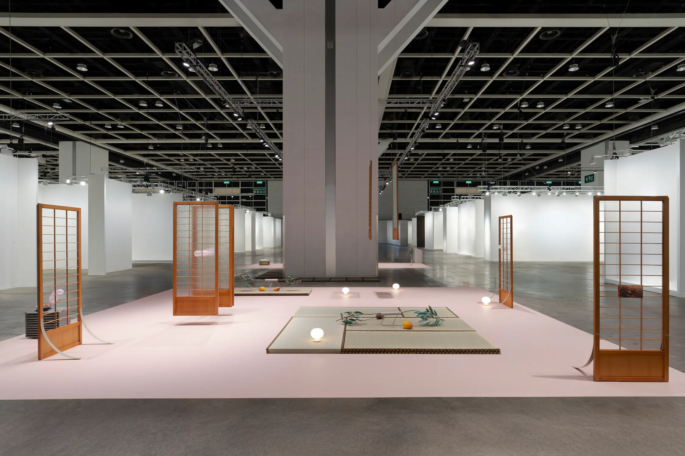 A modern art installation by Steph Huang features tatami mats, low tables, wooden shoji screens, a pink floor, scattered lamps, and objects beneath a large, industrial ceiling, set in a spacious gallery with white walls and exposed pipes.