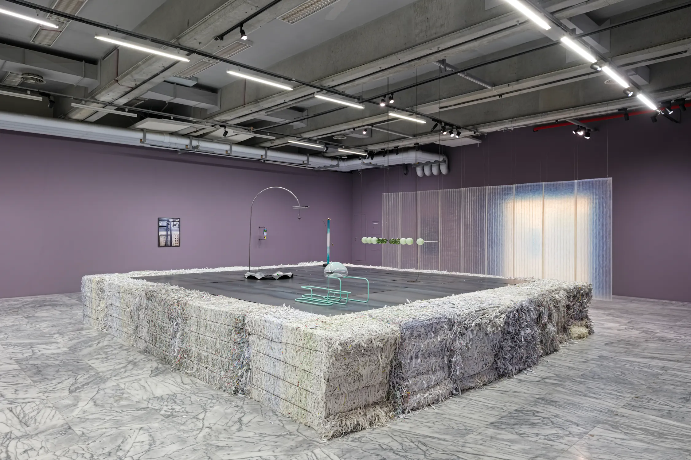 A modern art installation by Steph Huang in a gallery features a large rectangular pit bordered by bundled shredded paper. Inside, minimalist sculptures stand on a black floor, and a sheer, beaded curtain hangs along the purple wall at the back. The ceiling exposes pipes and lighting.