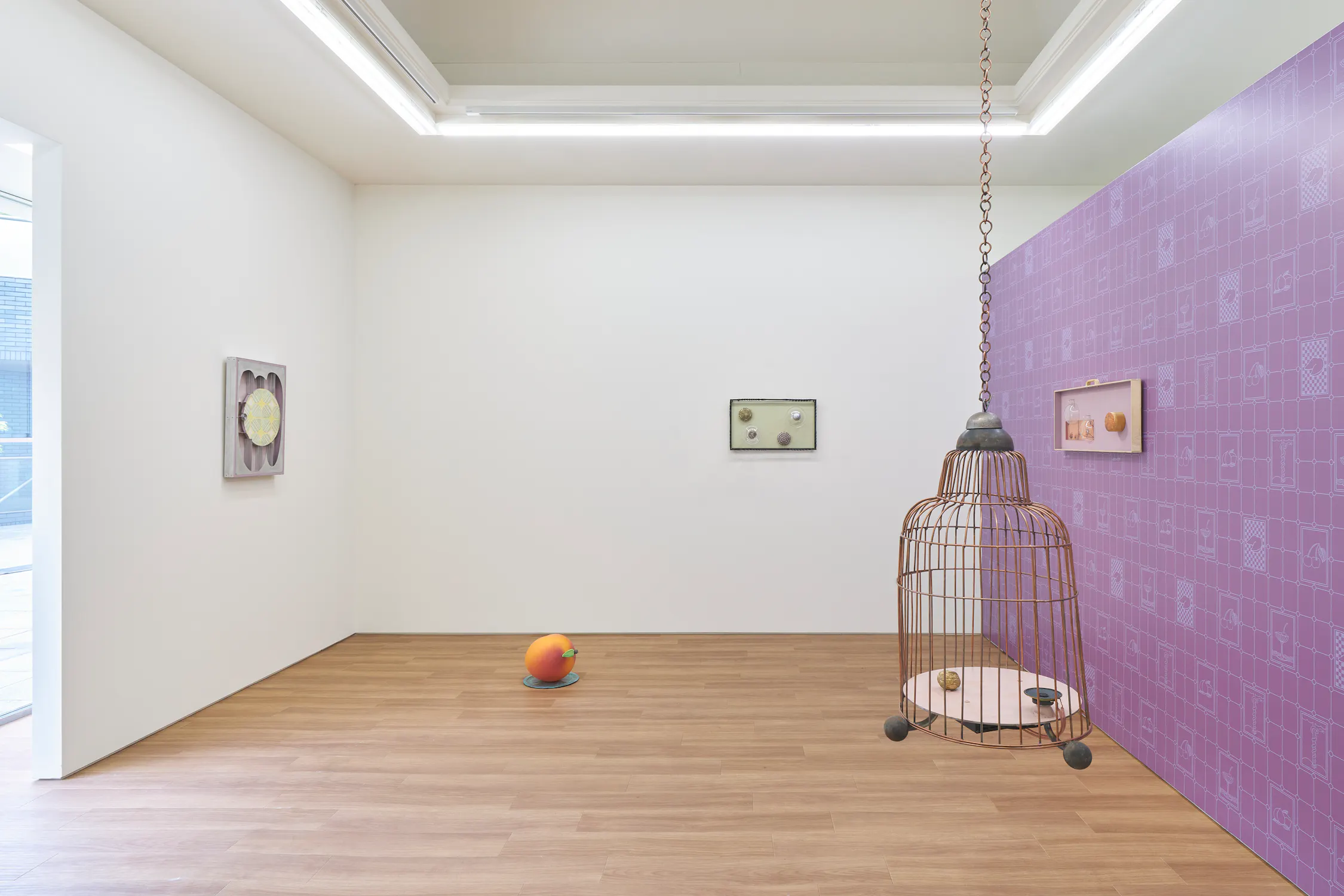 An installation by Steph Huang features white and purple walls, three framed artworks, a large birdcage hanging from the ceiling with objects inside, and a colourful spherical sculpture placed on the wooden floor. Light enters from a window on the left.