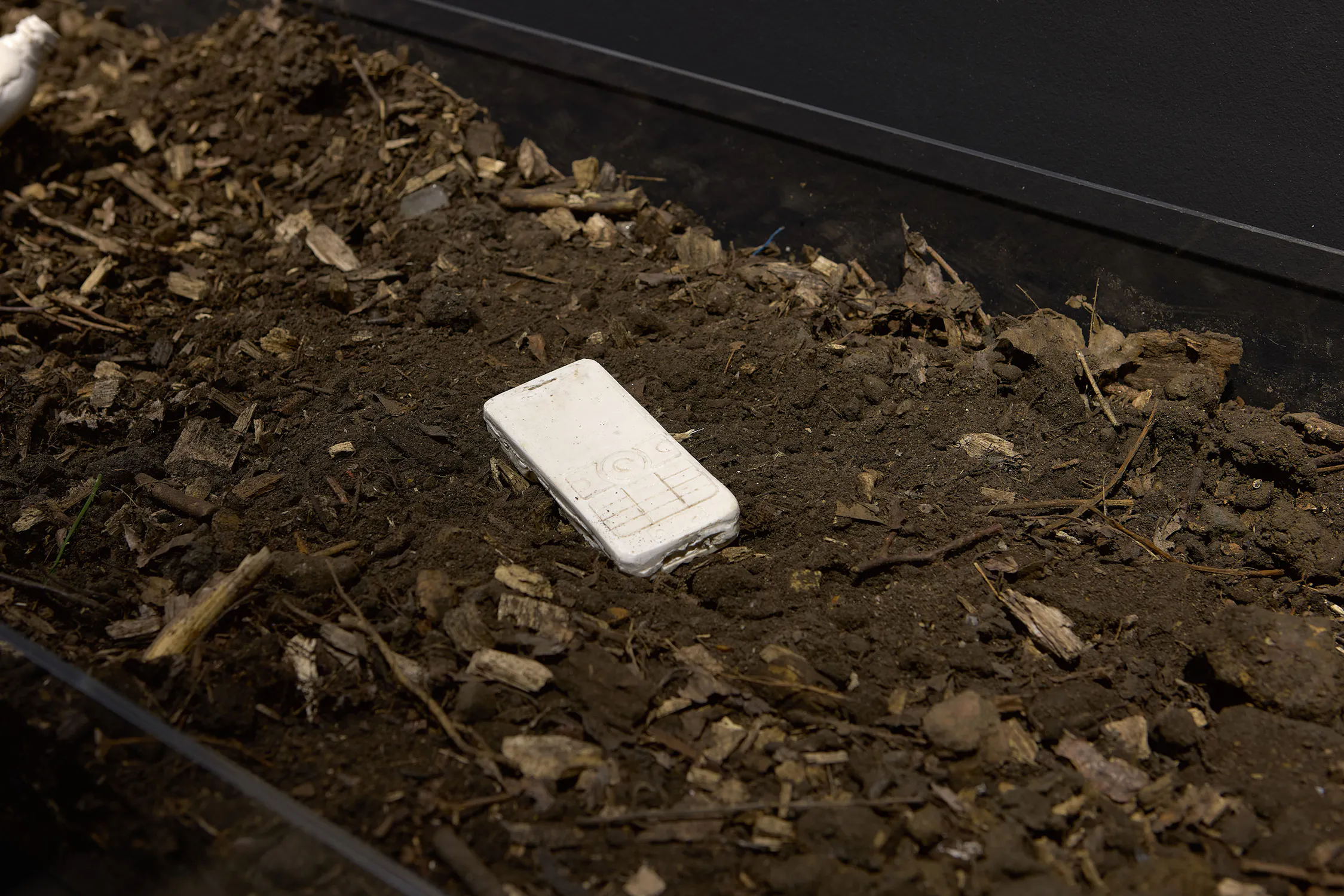 A white, rectangular object shaped like an old mobile phone is partially buried in dark, loose soil mixed with wood chippings and debris. The object appears slightly dirty and blends into the earthy background.