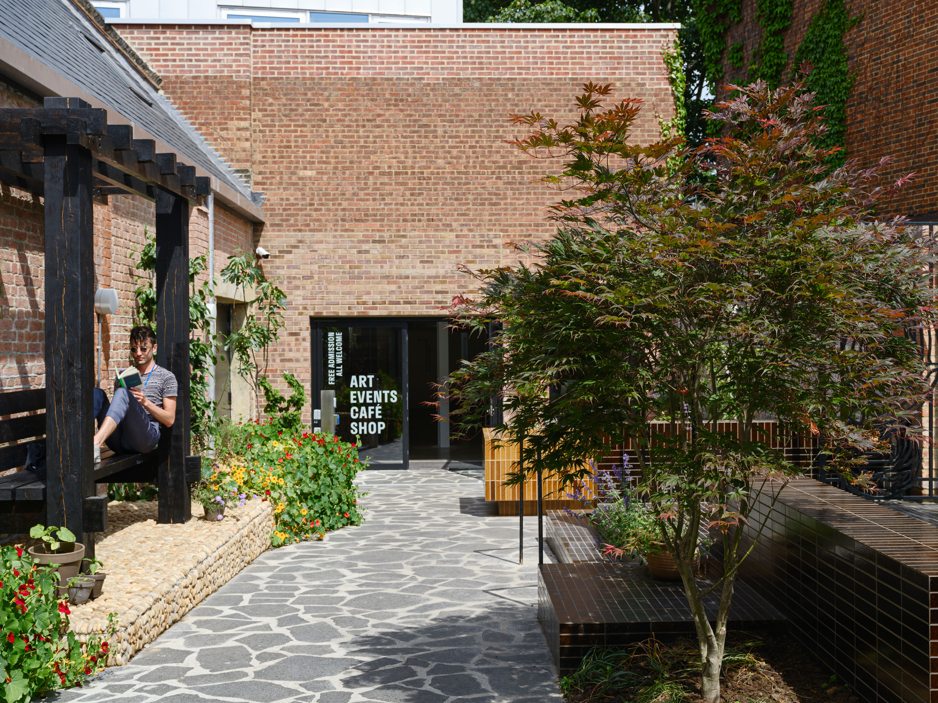A man sits and reads under a wooden pergola in a sunlit courtyard with stone flooring, lush plants, and a brick building. A sign on a glass door ahead lists: ART, EVENTS, CAFÉ, SHOP. A decorative tree and benches are on the right side.