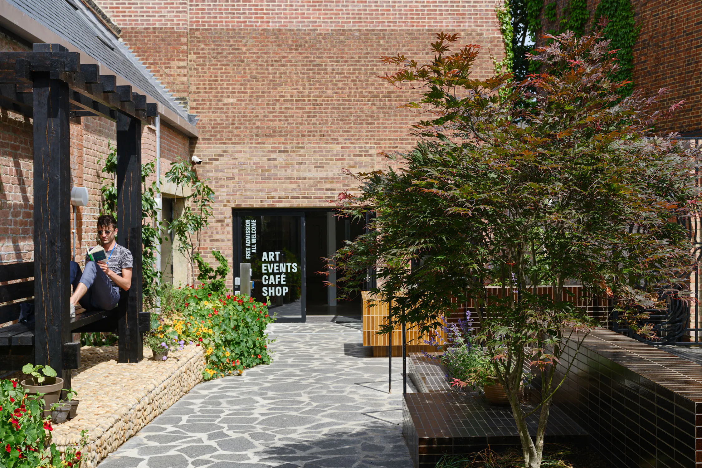 A man sits and reads under a wooden pergola in a sunlit courtyard with stone flooring, lush plants, and a brick building. A sign on a glass door ahead lists: ART, EVENTS, CAFÉ, SHOP. A decorative tree and benches are on the right side.
