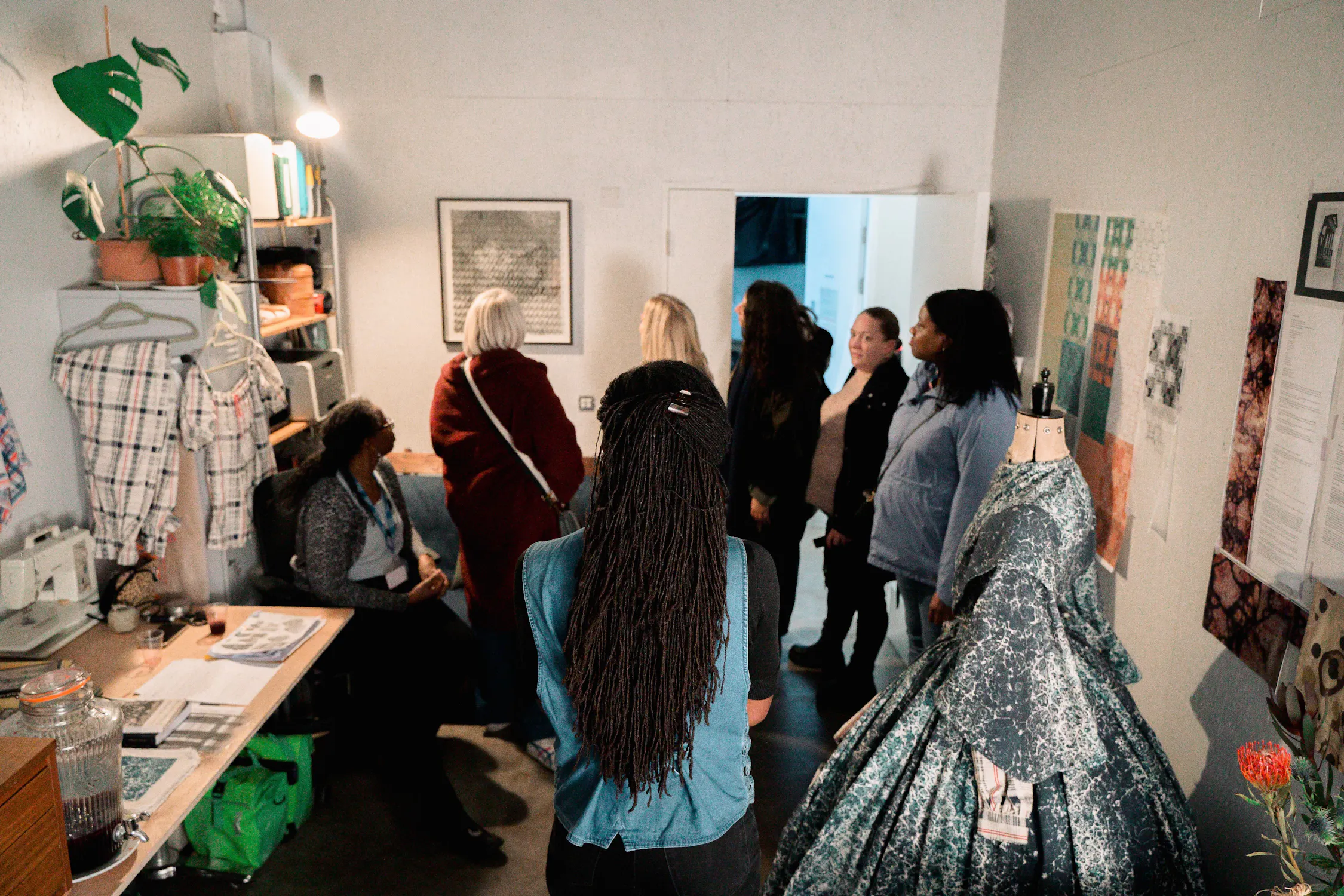 Inside Holly Graham's studio: a group of seven women gathers in a small, well-lit sewing studio. One woman sits at a desk with sewing supplies, while others stand nearby. A dress form with a blue patterned gown is on the right, and fabric samples and plants decorate the space.