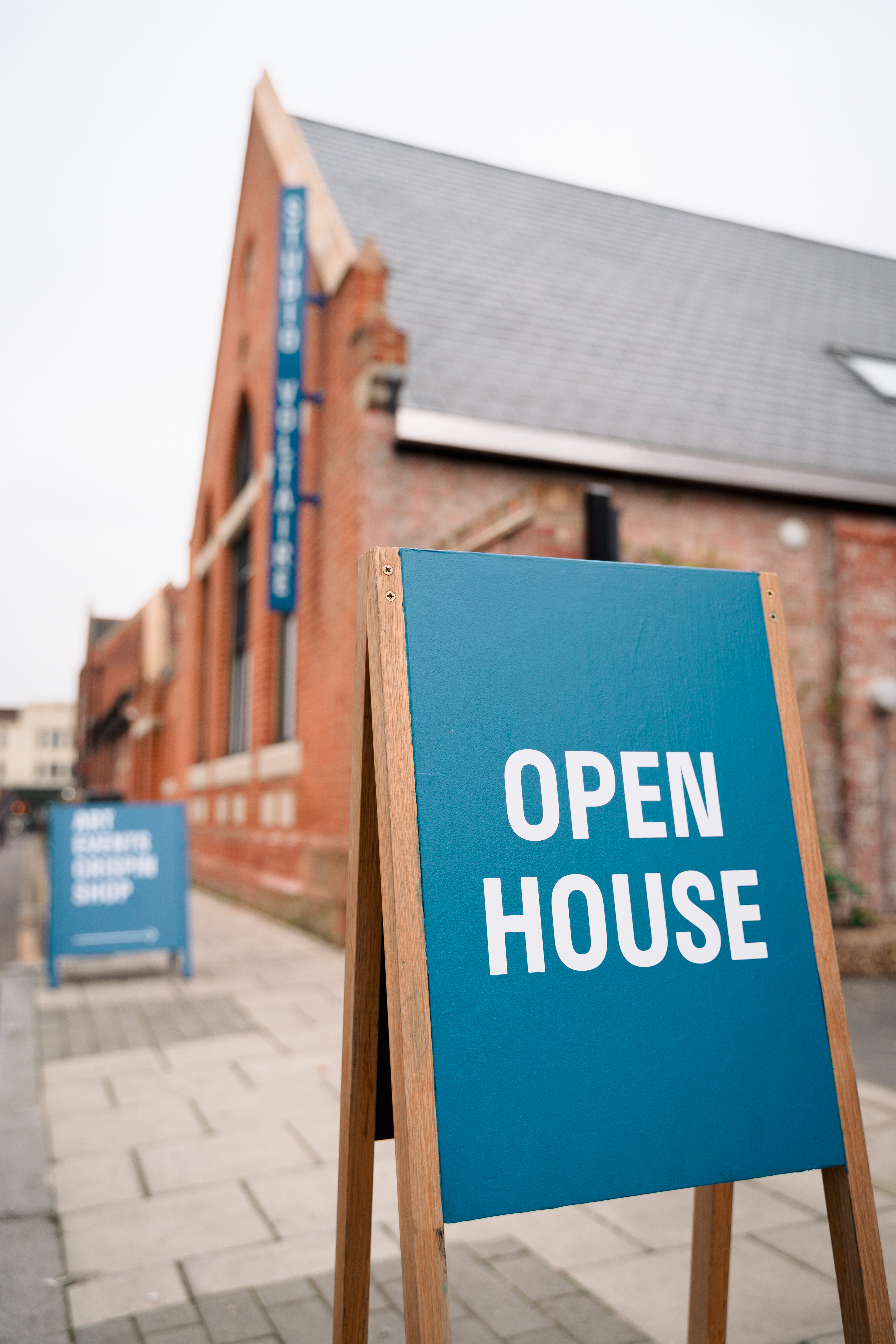 A blue A-board sign reads 'OPEN HOUSE' in white letters on a pavement outside a red-brick building with large windows and a sloping roof. Another similar sign is visible further down the walkway.