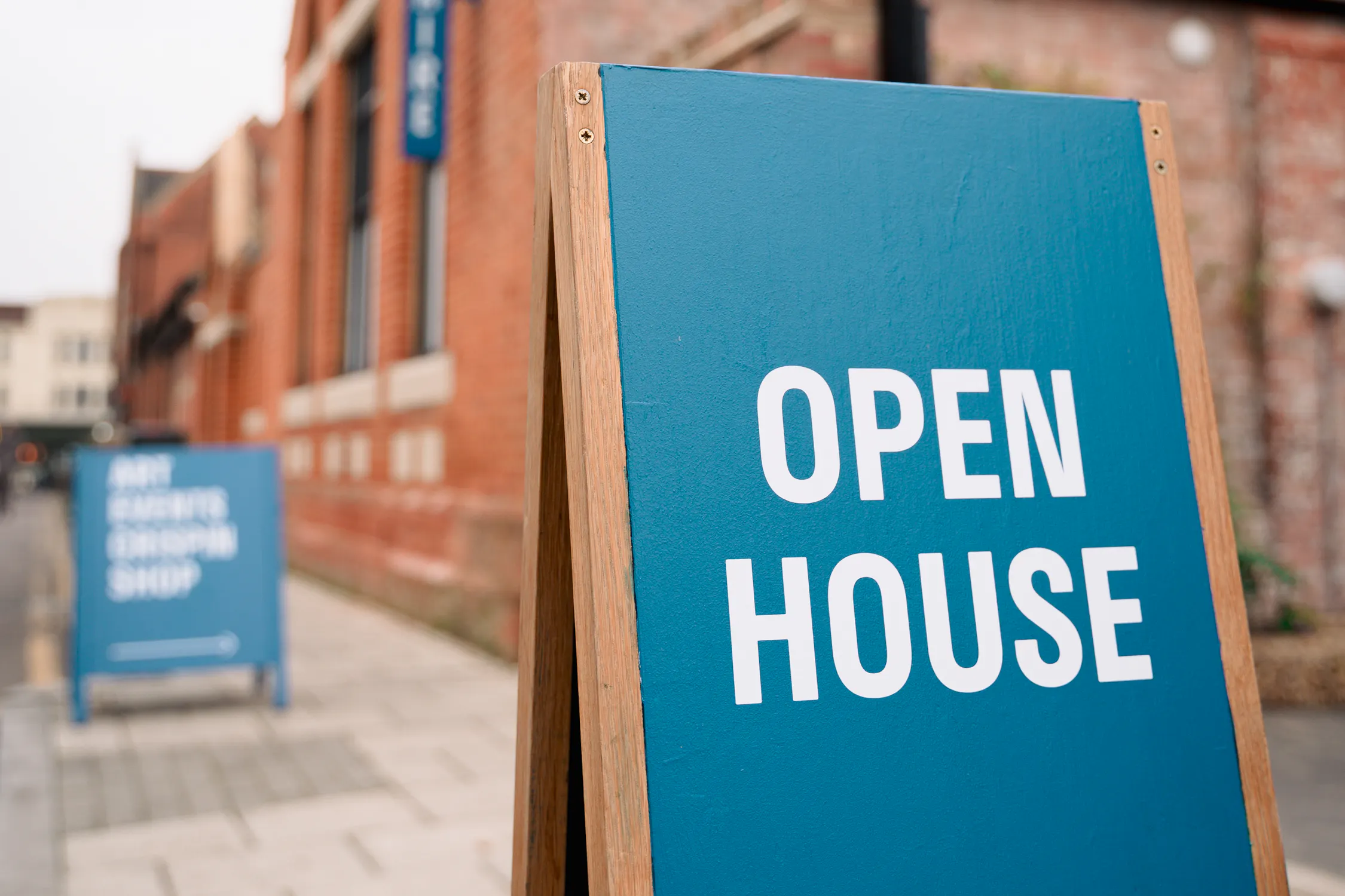 A blue A-board sign reads 'OPEN HOUSE' in white letters on a pavement outside a red-brick building with large windows and a sloping roof. Another similar sign is visible further down the walkway.