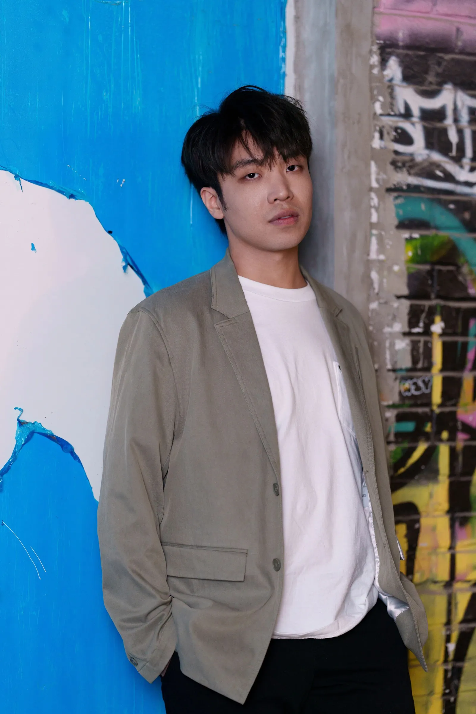 Jingzhou Yan hair wears a light grey blazer over a white T-shirt, standing with hands in pockets. He stands indoors against a bright blue wall and colourful, graffiti-covered brick. He gazes calmly at the camera.