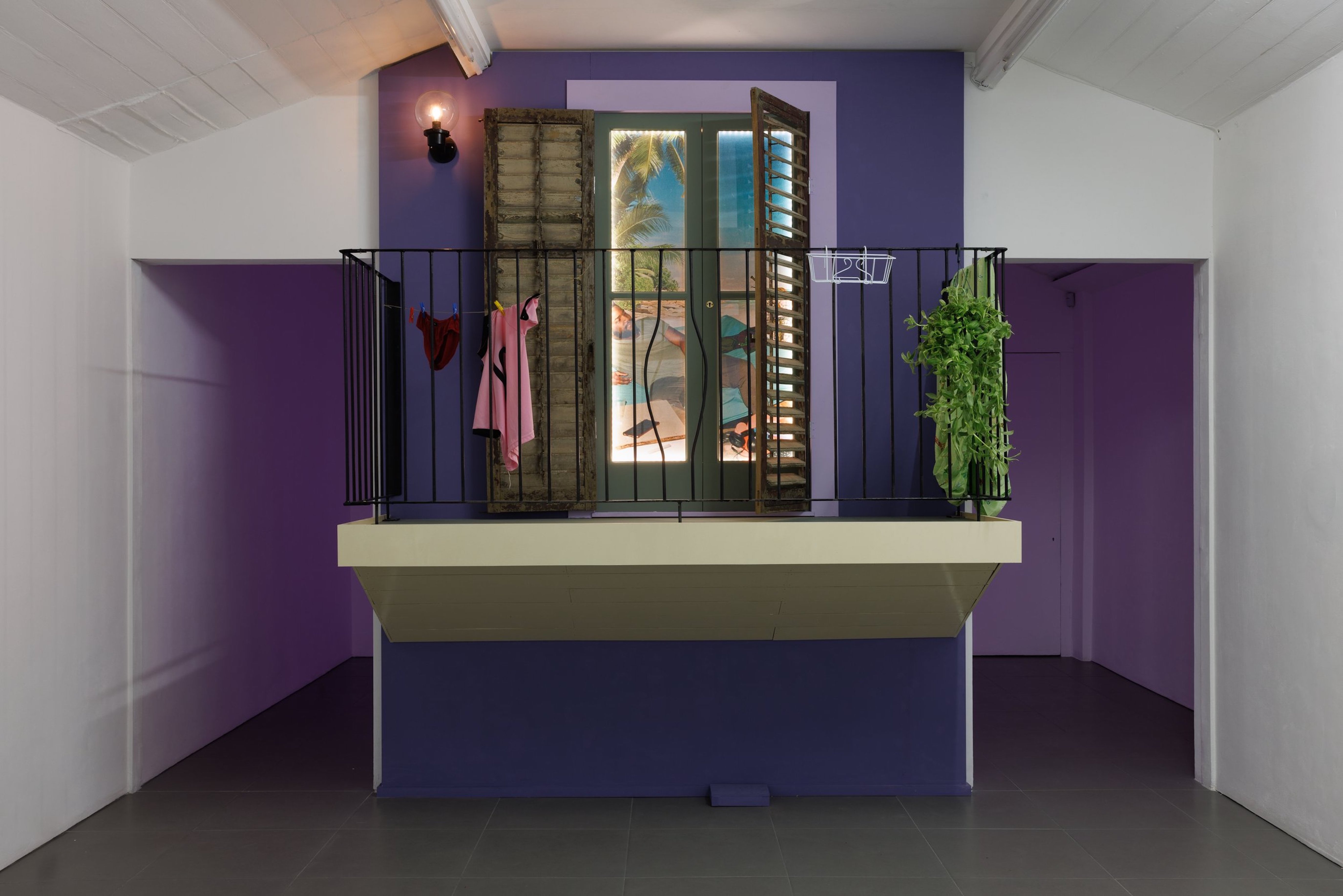 An installation by Bryan Giuseppi Rodriguez which features a small balcony with a metal railing features hanging clothes, a potted plant, and an open window with shutters. The wall is purple and a beach scene is visible through the window.