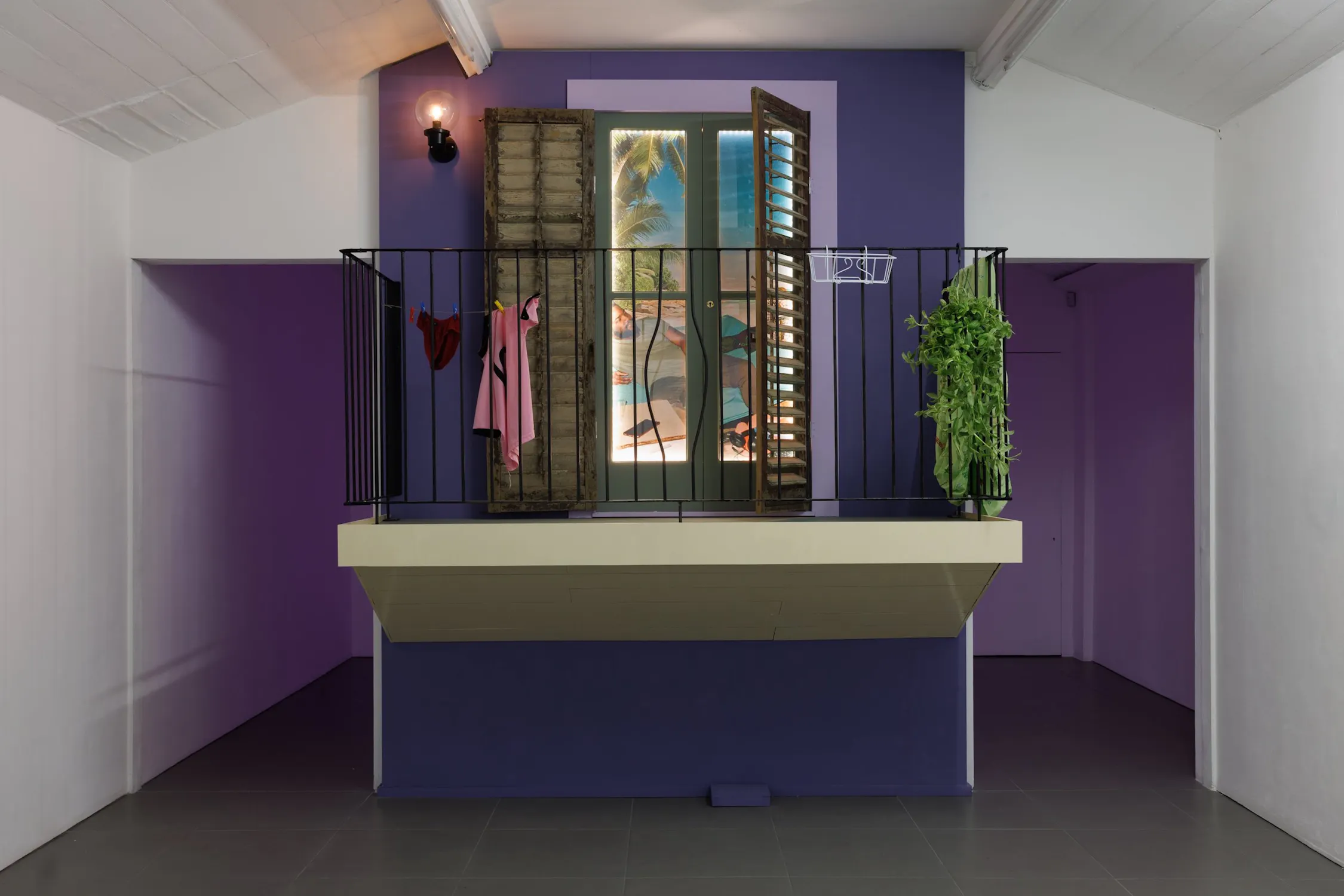 An installation by Bryan Giuseppi Rodriguez which features a small balcony with a metal railing features hanging clothes, a potted plant, and an open window with shutters. The wall is purple and a beach scene is visible through the window.