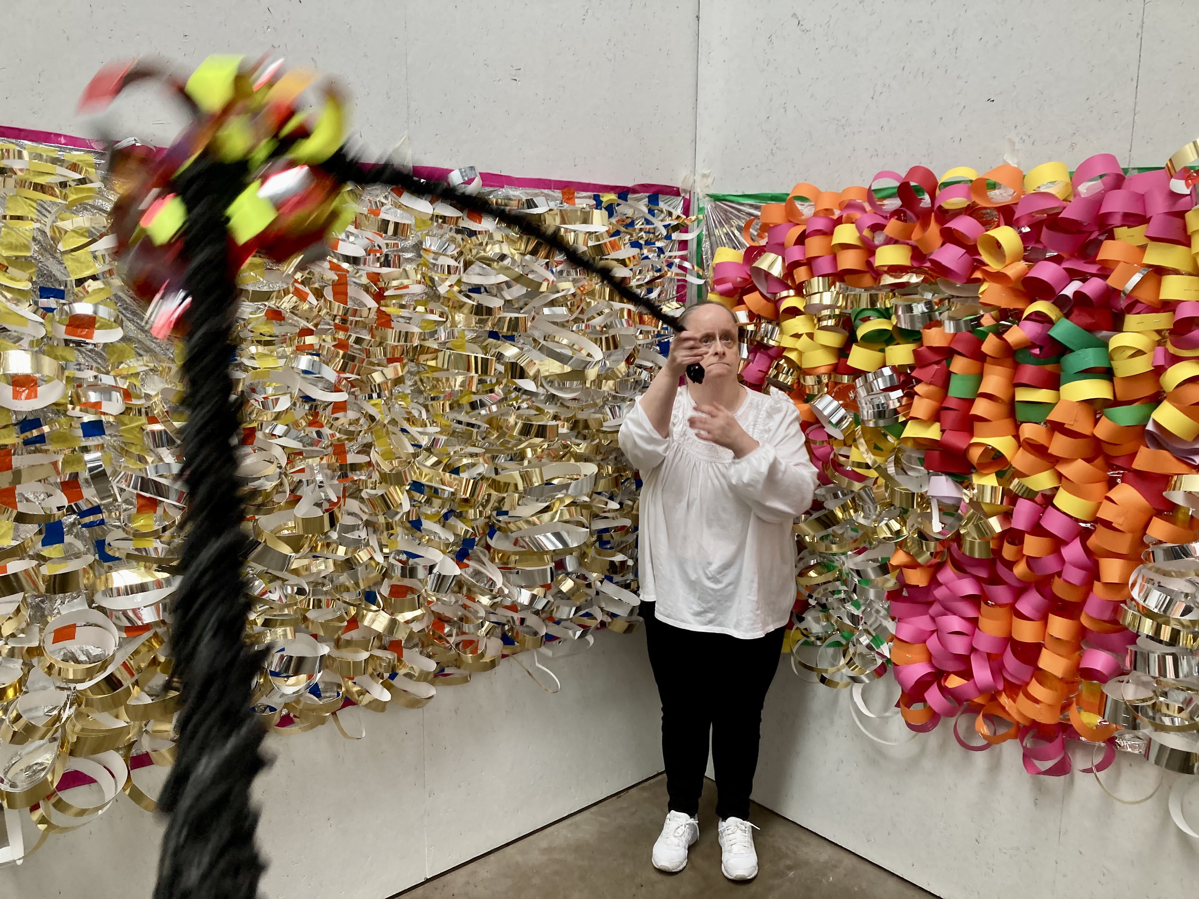 Linda Bell, wearing a white shirt and black trousers, stands in front of a wall decorated with shiny gold, silver, pink, and orange paper chains, taking a photo with their mobile. A blurred, colourful object swings in the foreground, partially obscuring the scene.