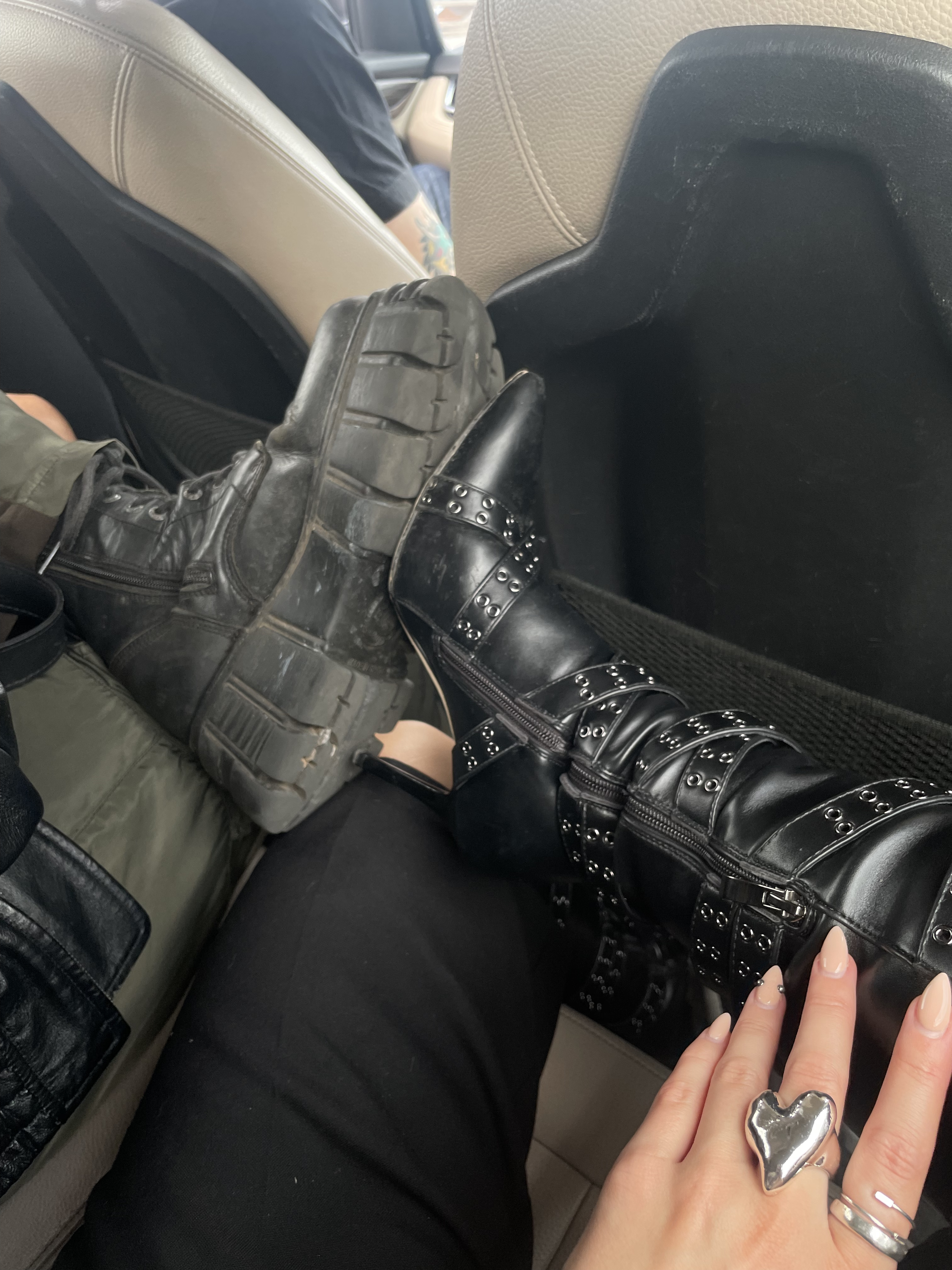 Two people sit in a car's back seat. One wears chunky black combat boots and green trousers; the other wears black studded high-heeled boots and a silver heart ring, with a manicured hand resting on their leg. Car seats and part of a window are visible.
