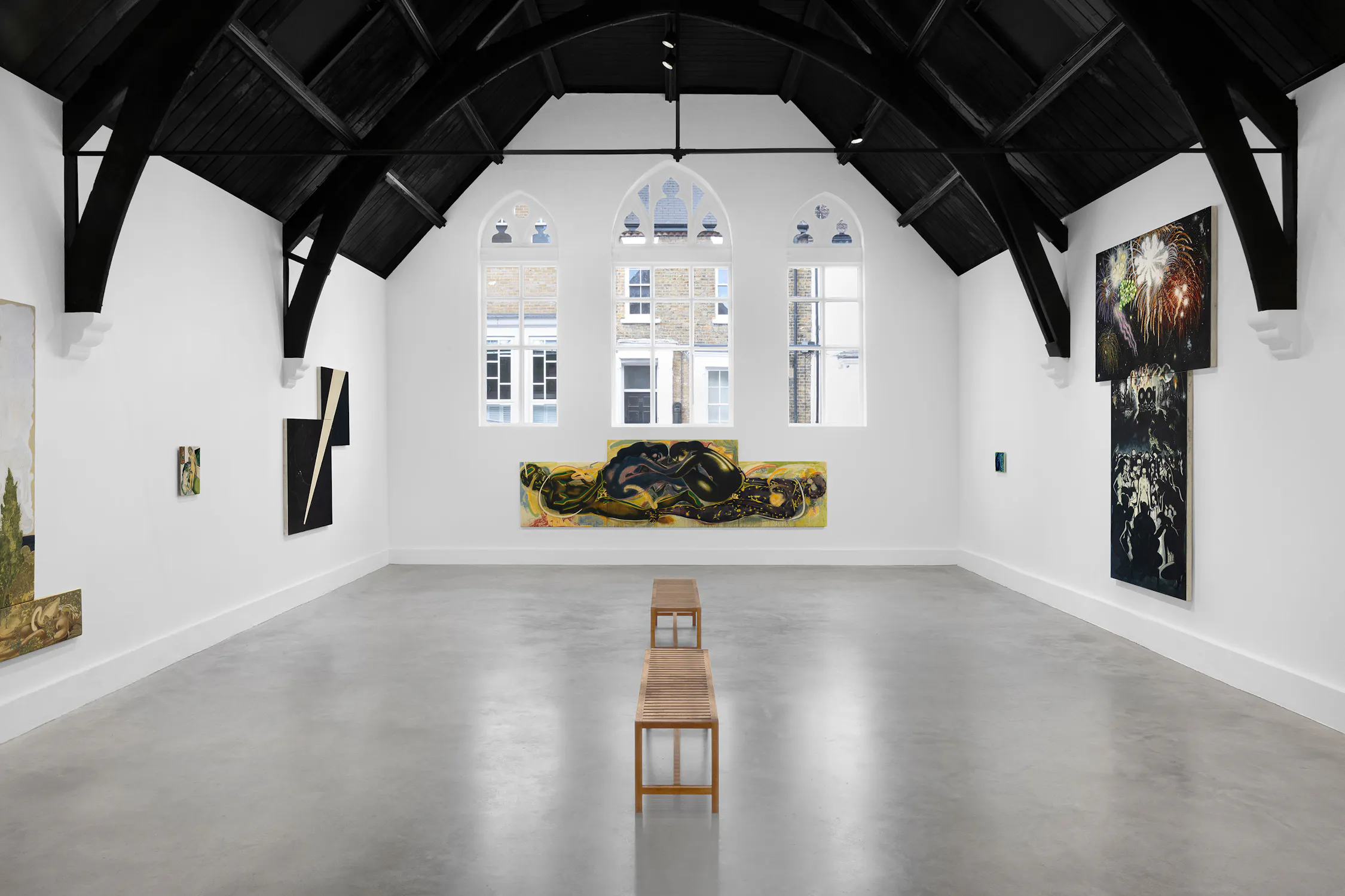 A bright art gallery with high, dark wood vaulted ceilings, arched windows, white walls, paintings by T. Venkanna on display, and two wooden benches centred on a grey polished floor. Natural light streams in from the large windows at the far end.
