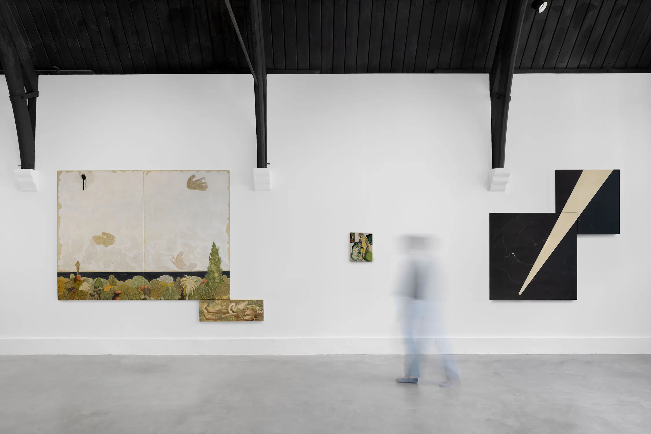 A blurred person walks past art by T. Venkanna: a large painting with birds and a landscape, a small central painting, and a black geometric artwork with a diagonal beige stripe. The ceiling is black with exposed beams. The floor is smooth and grey.