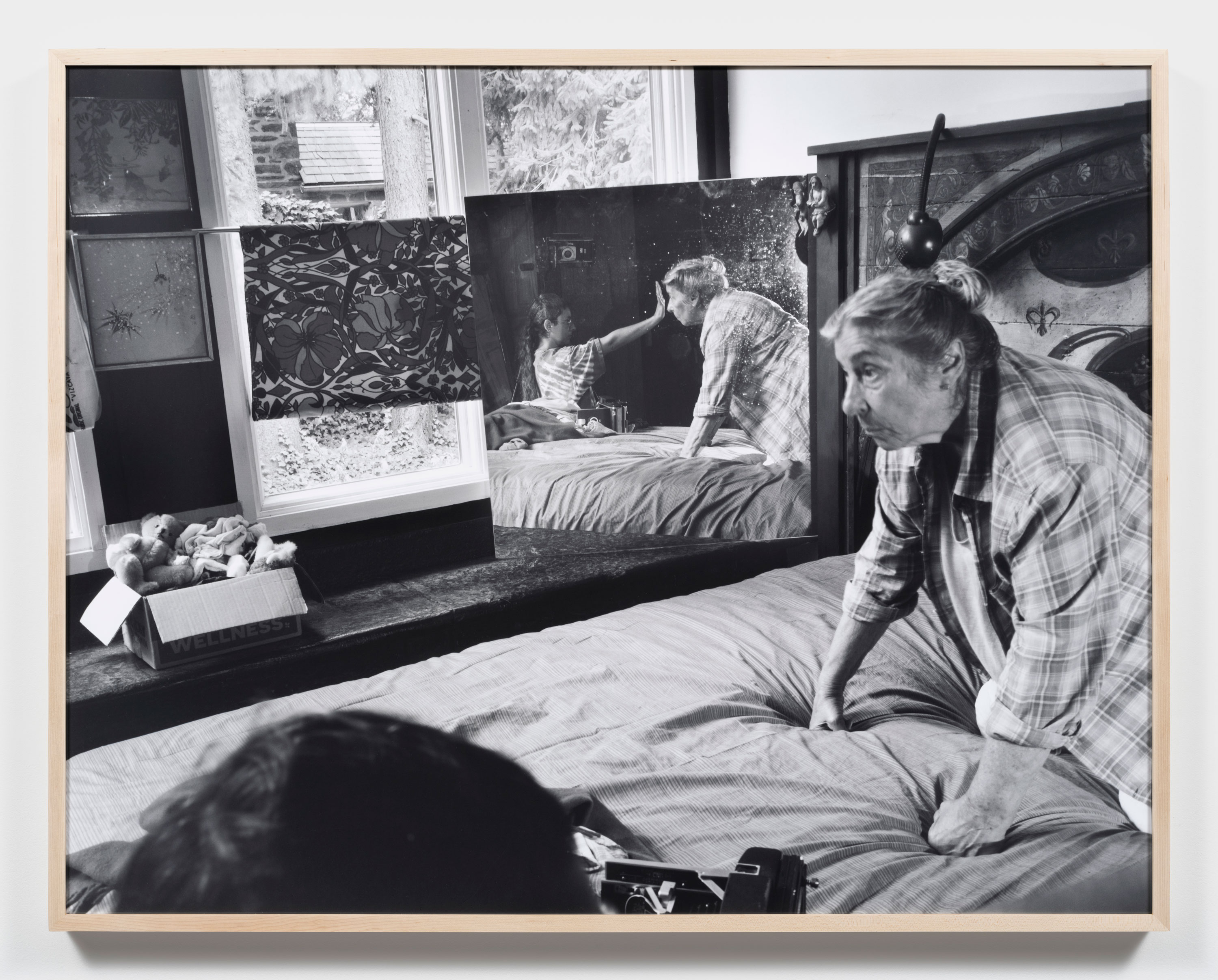 A black-and-white photo by Katherine Hubbard shows her elderly mother on a bed, leaning forward. A mirror by the window reflects them gently touching a child's face. The room has patterned curtains, a headboard, a stuffed animal box, and soft daylight from outside.