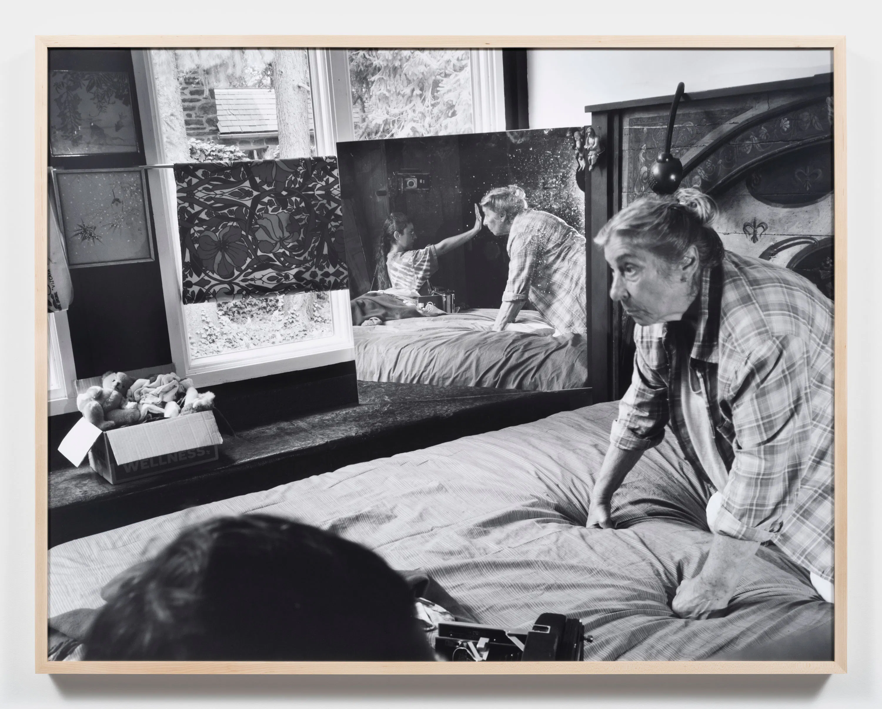 A black-and-white photo by Katherine Hubbard shows her elderly mother on a bed, leaning forward. A mirror by the window reflects them gently touching a child's face. The room has patterned curtains, a headboard, a stuffed animal box, and soft daylight from outside.