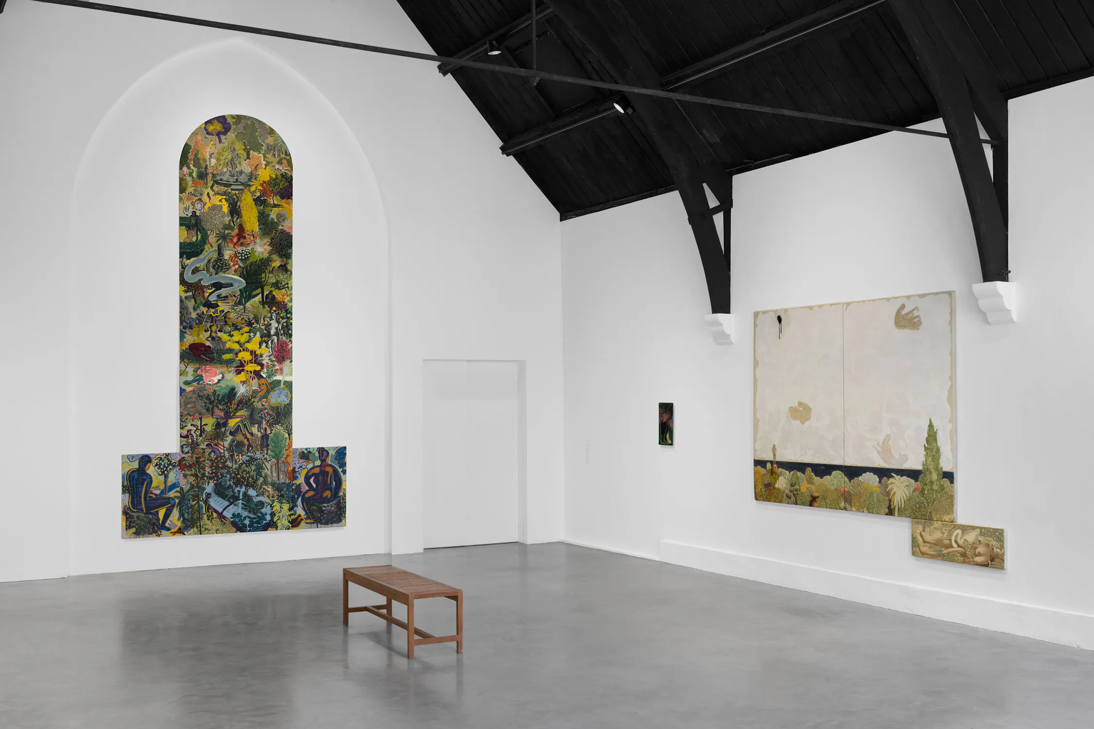 A minimal gallery room with white walls, grey floor, and dark vaulted ceiling. Abstract colourful paintings hang on walls; one is large and arch-shaped, the other is rectangular. A simple wooden bench sits at the centre of the room.