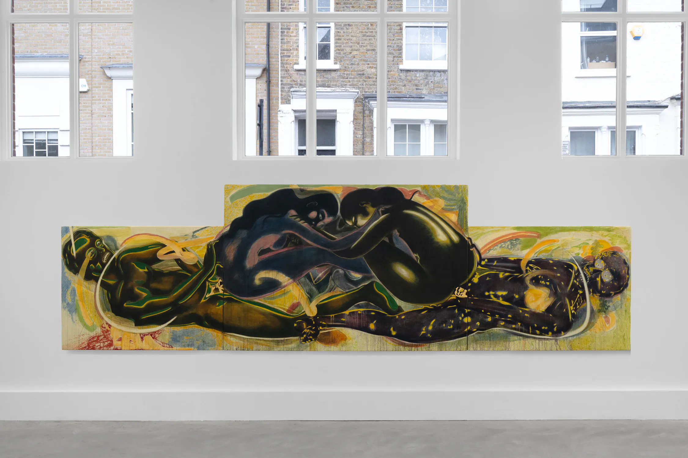 A large, colourful painting by T. Venkanna hangs on a white gallery wall beneath tall windows. The artwork features two abstract, intertwined figures with dark skin tones and dynamic yellow, green, and blue brushstrokes on a tiered canvas. The background is vibrant and swirling.