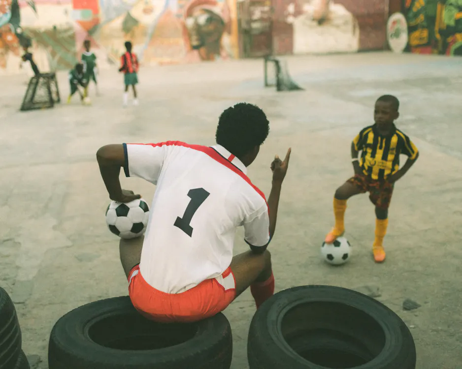 Photograph by Carlos Idun-Tawiah. A person in a white kit with a black "1" squats on tyres, holding a football, and gestures as they talk to a young boy in a yellow-and-black striped kit holding a football. Other children play football in the background on a concrete court with mural-covered walls.