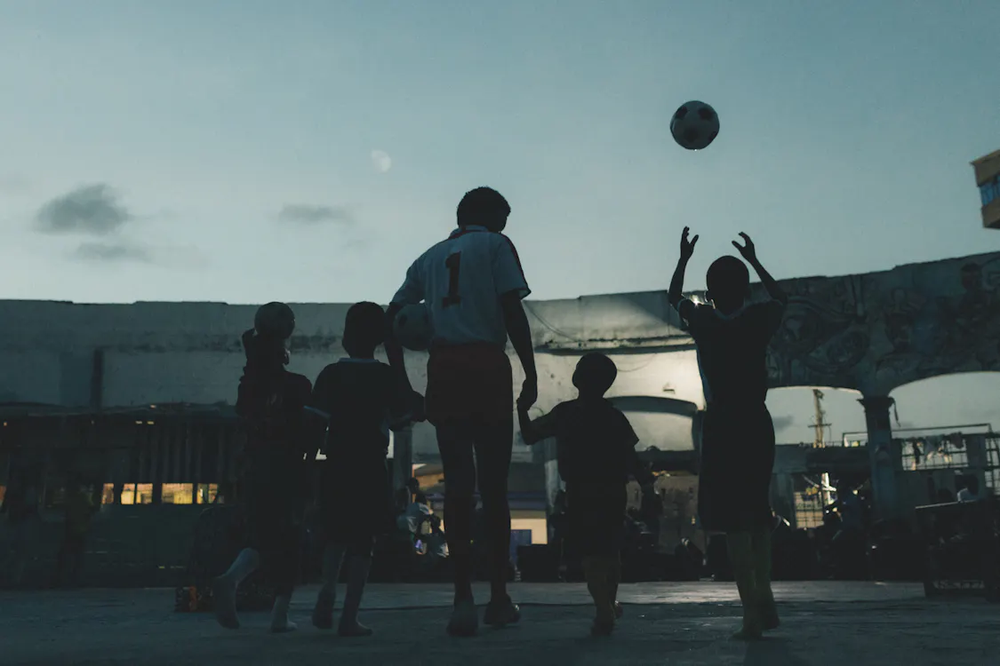 Photography by Carlos Idun-Tawiah. A group of children play football at dusk on a concrete lot. Their silhouettes stand out against the dim sky. One child wears a shirt with the number 1, another tosses a football in the air. Buildings and graffiti are visible in the background.