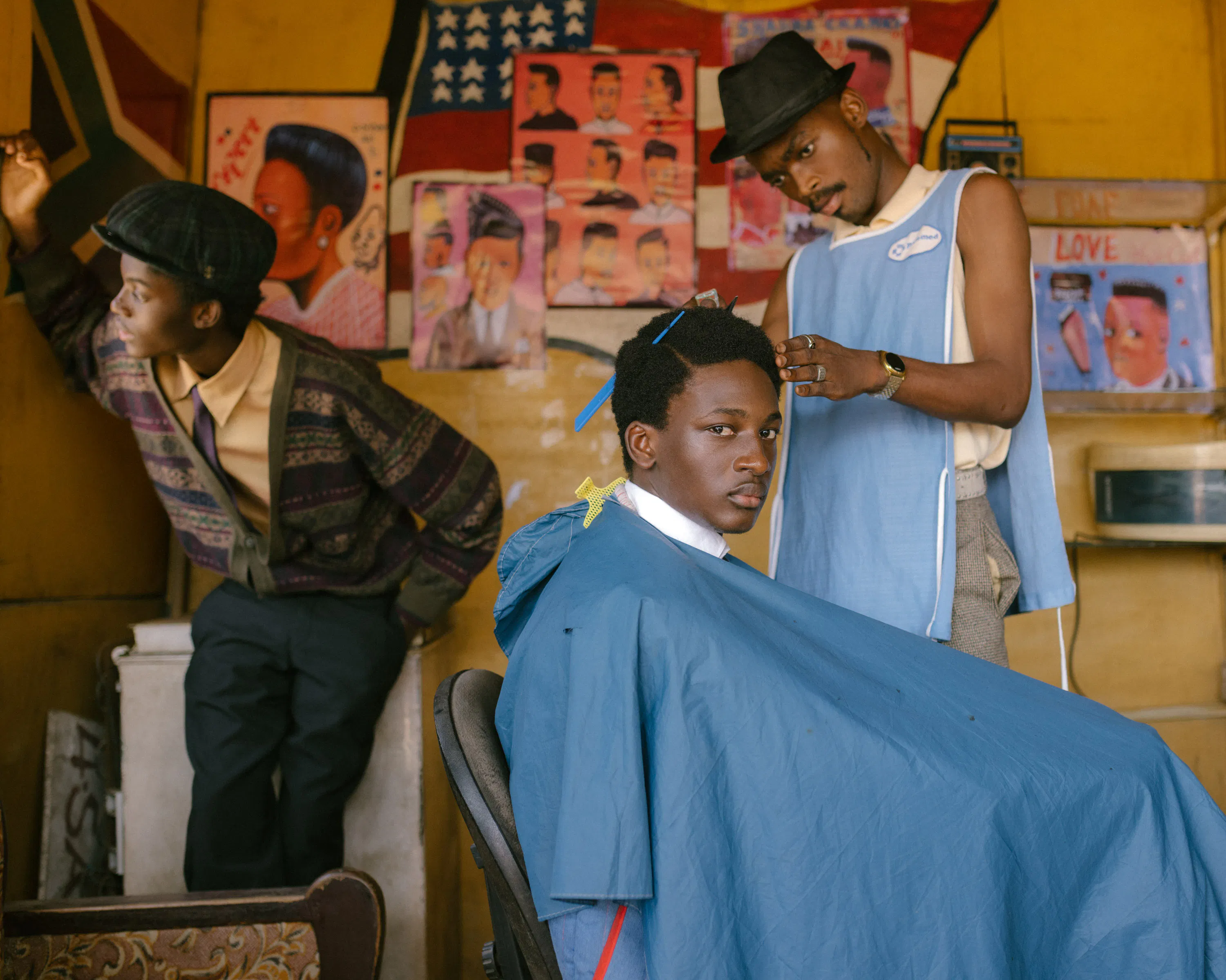 Photography by Carlos Idun-Tawiah. A young man sits in a barber’s chair covered with a blue gown, looking at the camera. A barber in a blue apron trims his hair. Another man in a checked cap leans against the wall. Colourful hairstyle posters and a US flag decor adorn the yellow-walled barber’s shop.