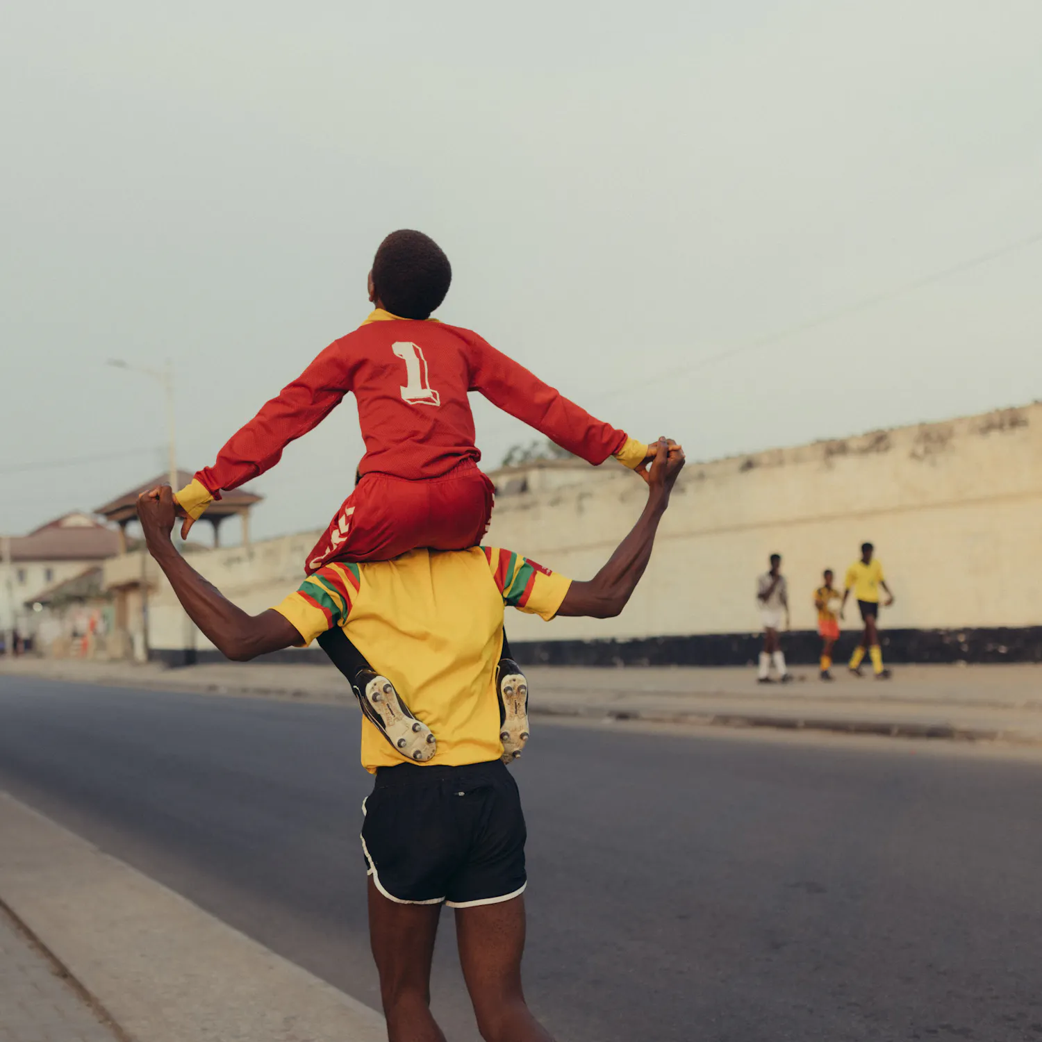 Photography by Carlos Idun-Tawiah. A young child in a red outfit with the number 1 sits on an adult’s shoulders, arms outstretched, holding the adult’s hands. The adult wears a yellow shirt and black shorts as they walk along a quiet street. Other people and a building are visible in the background.