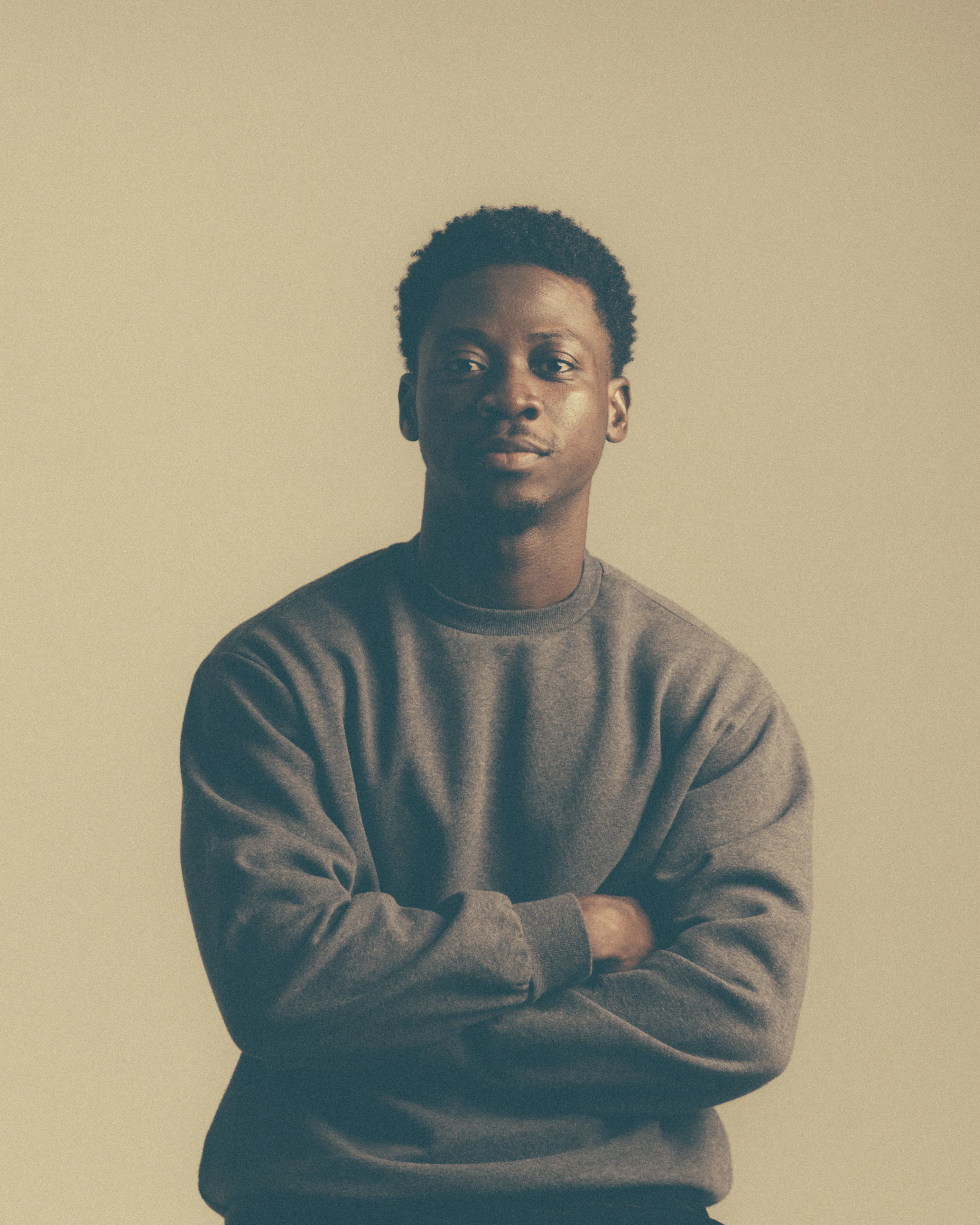 Carlos Idun-Tawiah. He has short curly hair and wears a plain grey sweatshirt. He stands against a beige background with his arms crossed and a neutral expression, looking directly at the camera. The lighting is soft, giving the image a calm and muted tone.