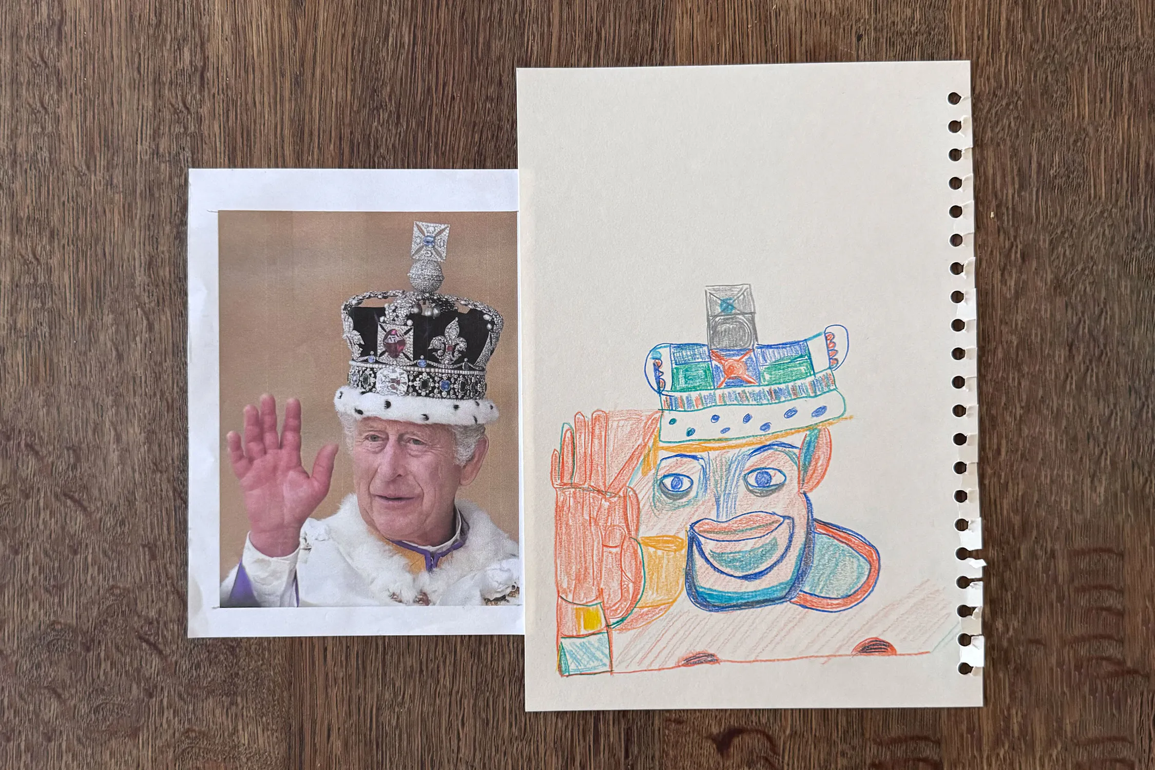 A printed photo of King Charles III in a royal crown and robe raising his hand is placed next to a colourful pencil drawing by Christine Hazell, mimicking the photo, featuring the same gesture and crown, both on a wooden surface.