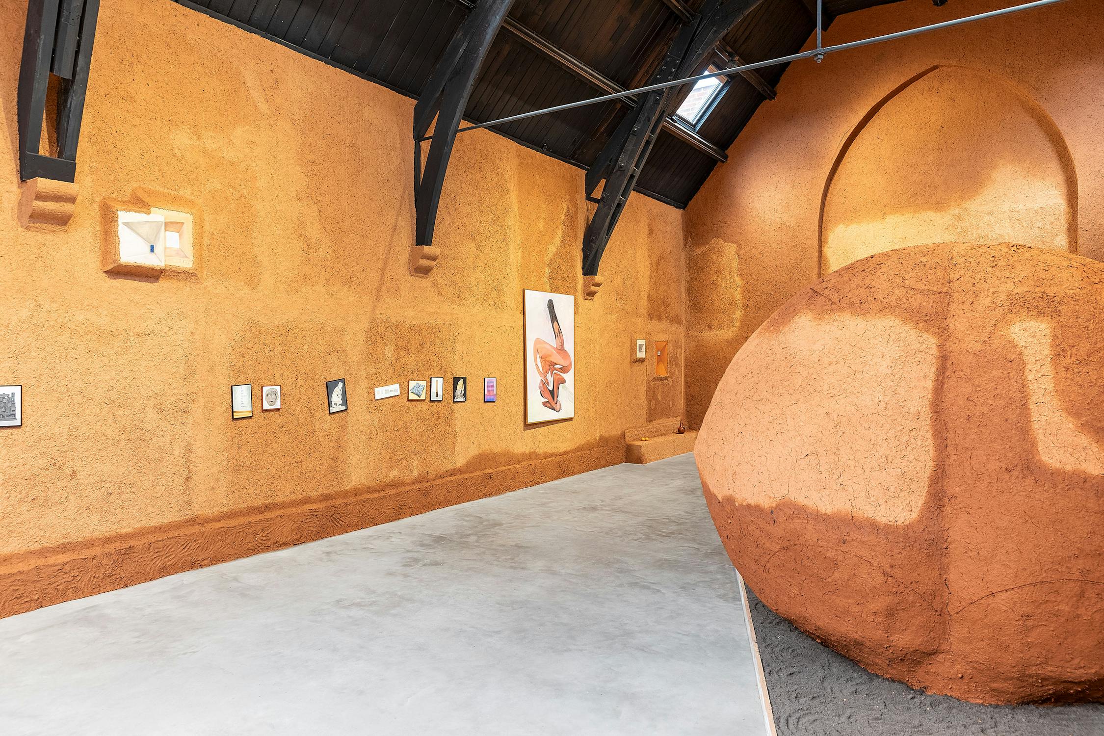 A gallery with walls entirely covered in terracotta coloured mud clay, with a vaulted black ceiling. In the centre of a room stands a large round sculpture made of mud clay, sitting in a raised bed of brown earth. Small paintings of publicly held artworks are arranged on the walls