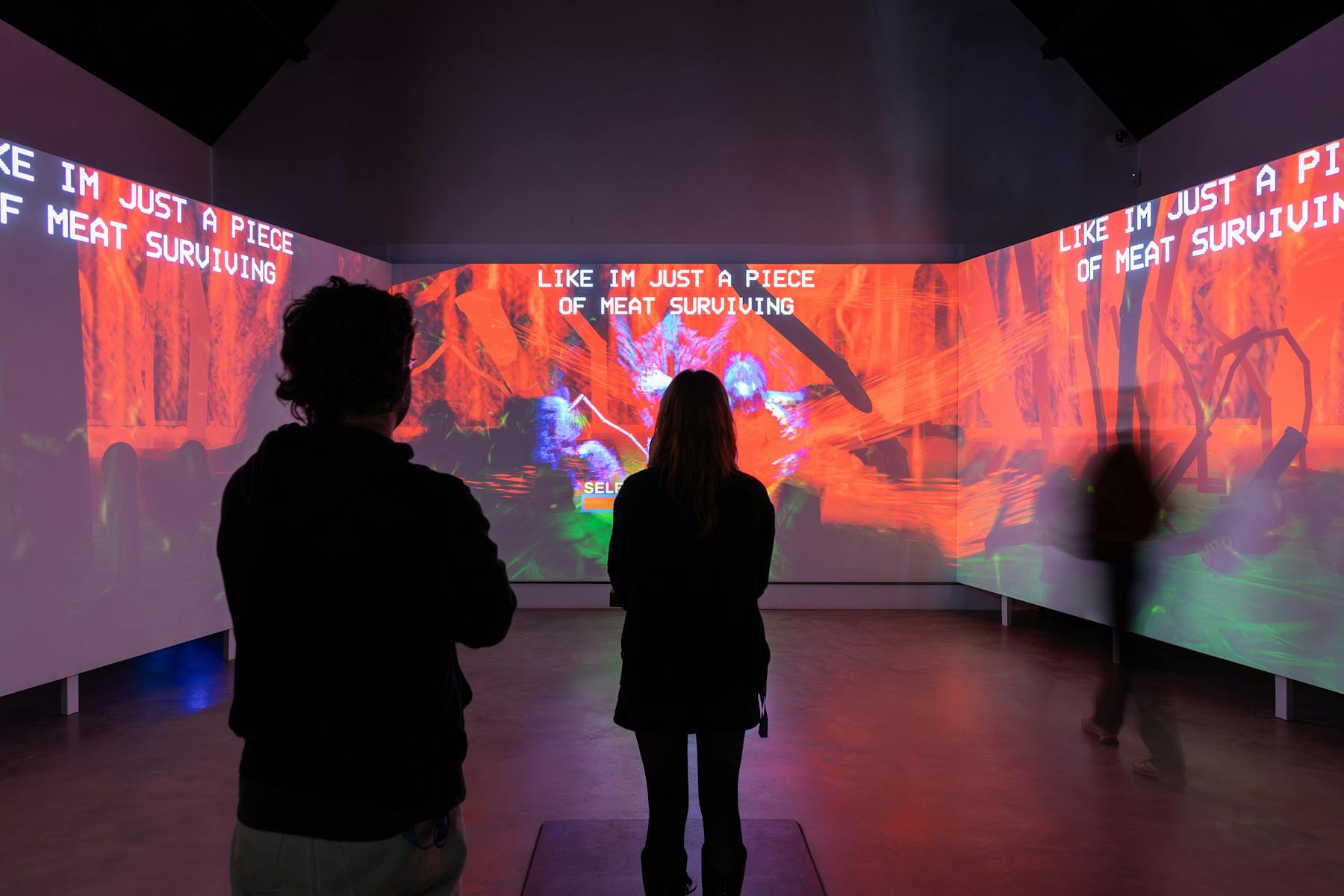 a photograph of three large wall-sized screen with colourful projects. There are three blurry figures in the foreground.Text on the screens reads 'LIKE IM JUST A PIECE OF MEAT SURVIVING'