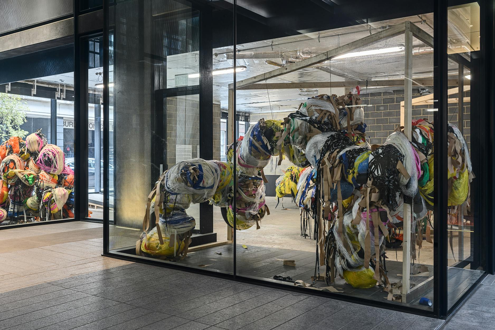 A large, abstract sculpture made of colourful, twisted fabric and materials is displayed inside a glass-walled gallery. The installation forms tangled, looping shapes. The surrounding space is industrial, with brick walls and exposed ceilings.