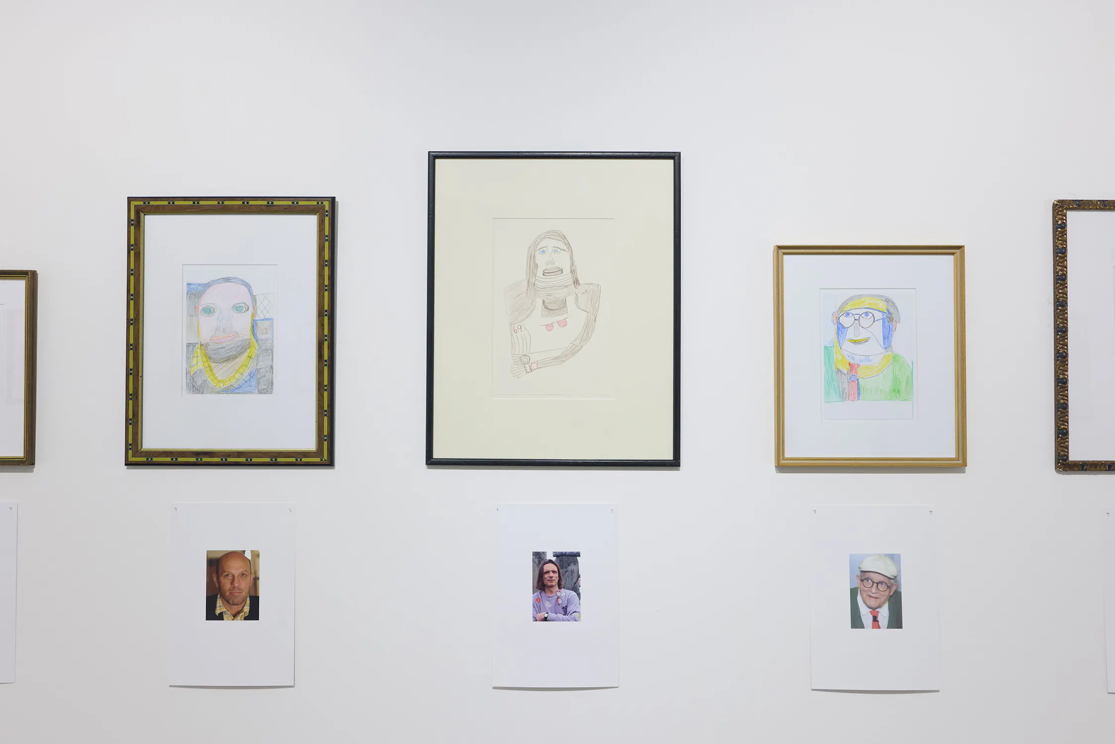 Three drawn portraits in ornate frames hang on a white wall. Directly below each are reference photos used by the artist to create the drawings. The centre portrait is pencil-drawn, while the others are colourful. No visible museum visitors.