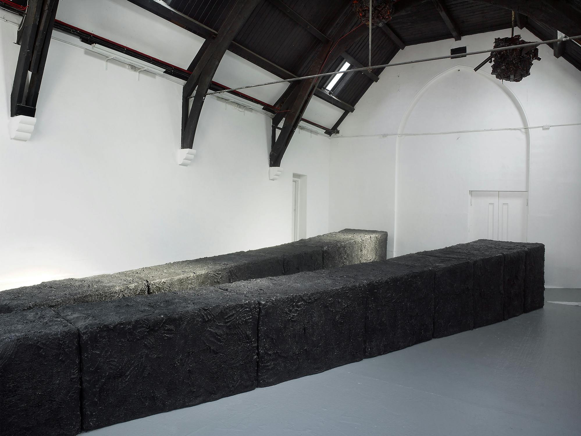 A minimalist art installation features large, rectangular black blocks arranged in a zigzag formation on a light grey floor inside a white, high-ceilinged gallery with arched beams and overhead lighting. The blocks have a textured, rough surface.