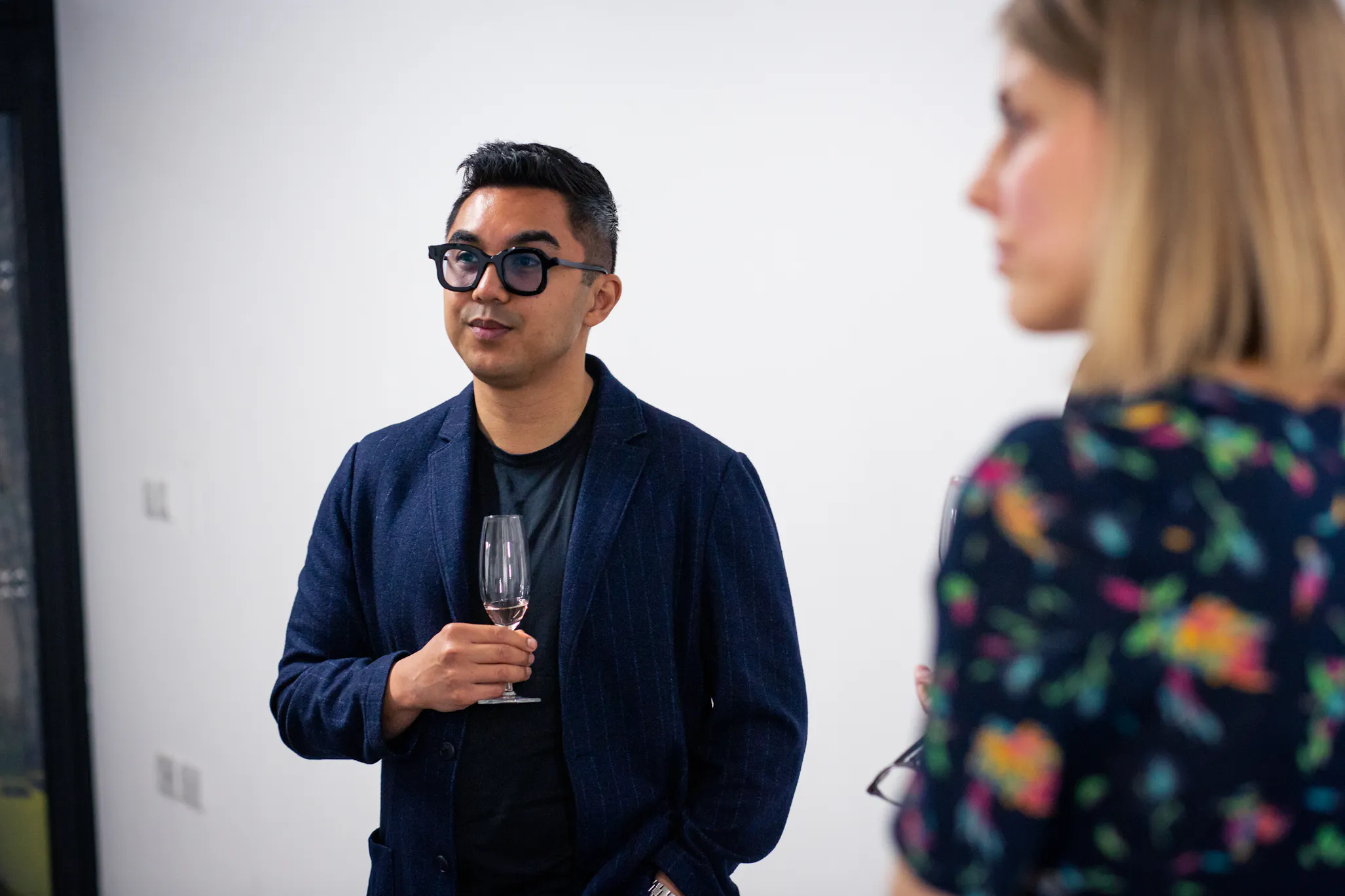 A man in dark glasses and a navy blazer holds a wine glass, standing in front of a white wall. He looks thoughtfully ahead. A woman with blonde hair, wearing a floral dress, is blurred in the foreground on the right side of the photo.