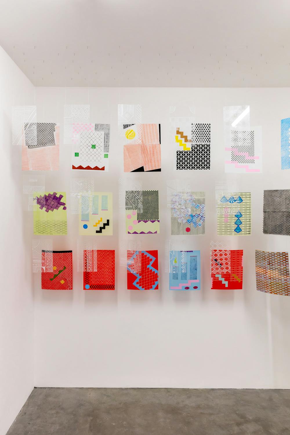 A wall displays an art installation featuring rows of colourful, abstract designs. Each piece has a mix of geometric shapes, lines, and patterns, hanging individually on transparent sheets against the white wall, creating a vibrant and modern look.