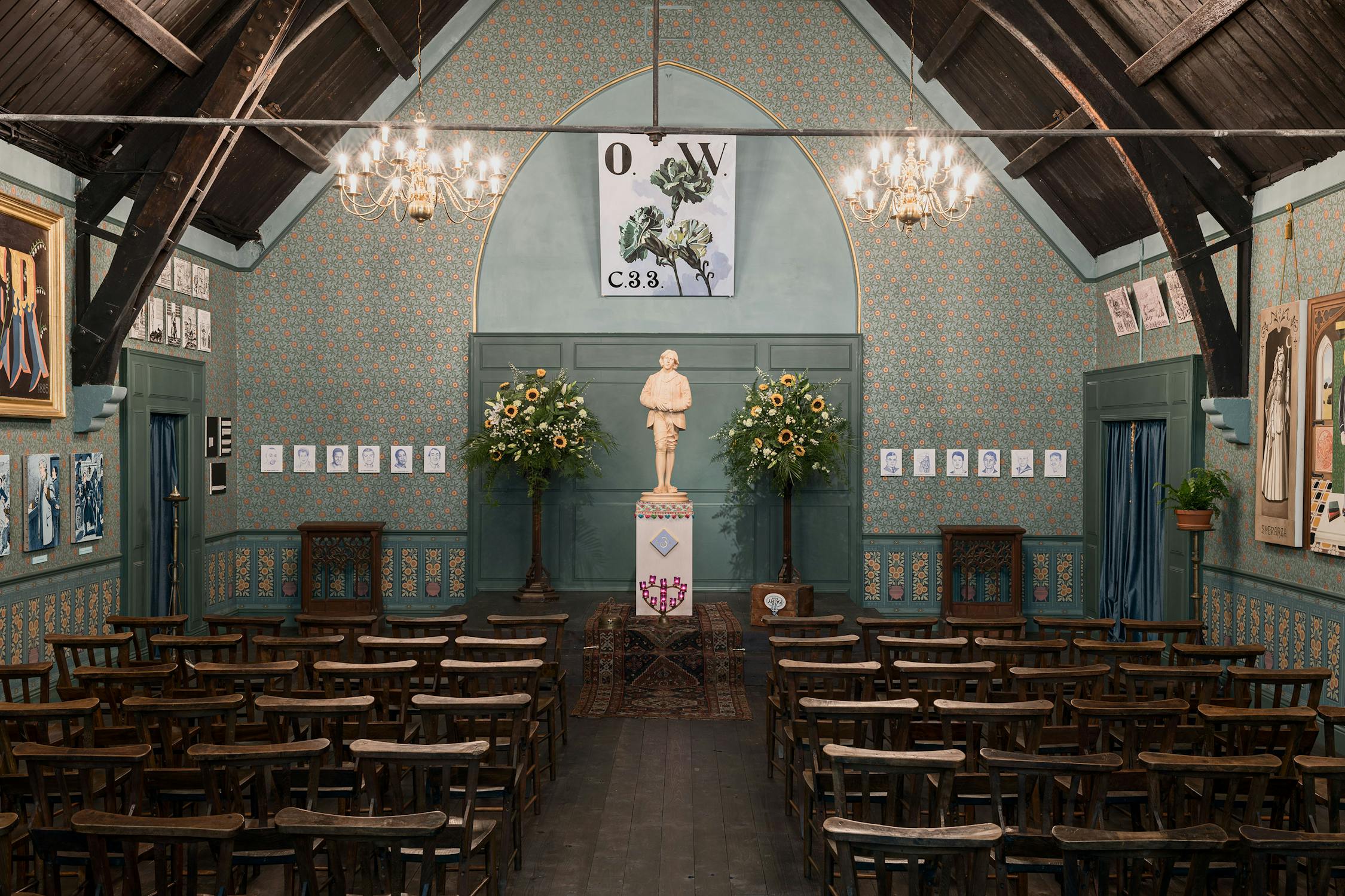 A Victorian-era hall with wooden chairs facing a central statue on a pedestal, flanked by tall floral arrangements. The room has patterned wallpaper, chandeliers, and framed art. Above the statue, a banner reads "O.W. C.S.3." under an archway.