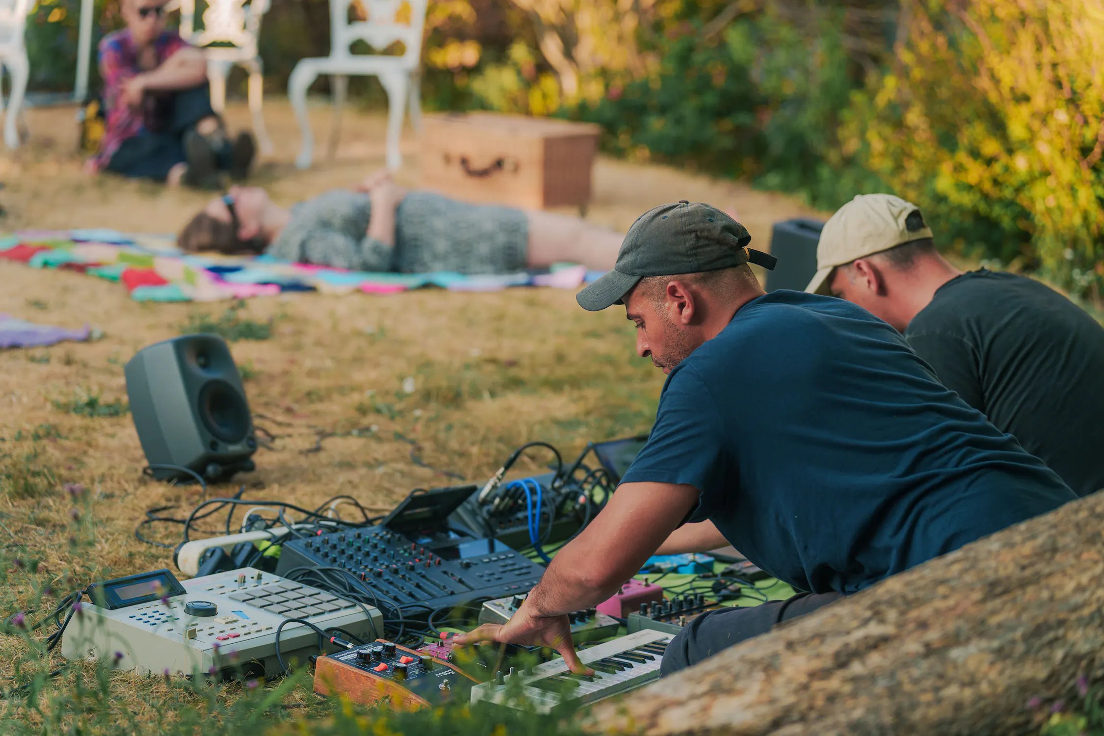 Abbas Zahedi and Tom Harris sit on the grass outdoors, manipulating a collection of synthesisers, mixers, and audio equipment. In the background, a person rests on a colourful blanket while another sits on a chair.