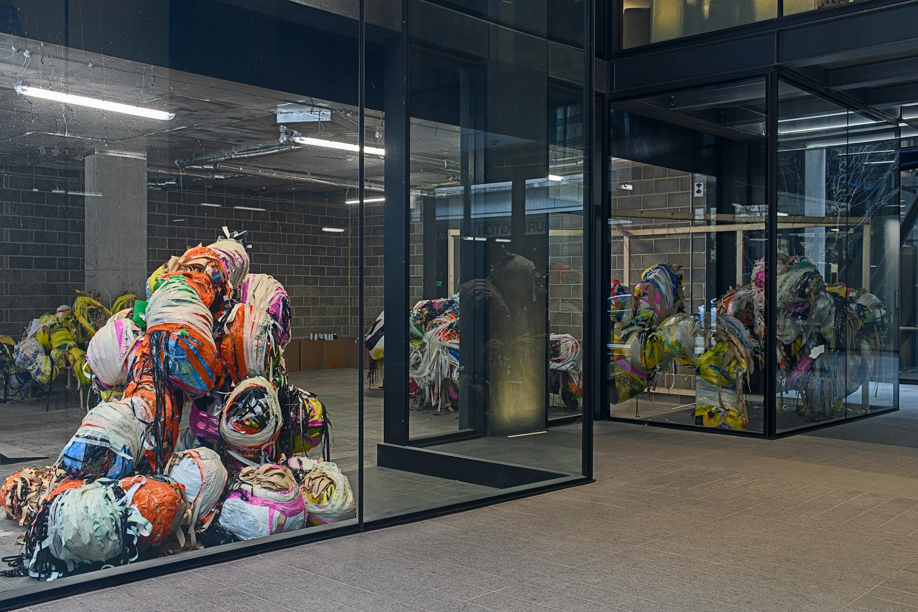 A modern building with floor-to-ceiling glass walls displays piles of colourful, abstract, bundled materials or fabric inside. The reflection in the windows shows more of these sculptural bundles filling the interior space. The exterior is paved and minimalistic.