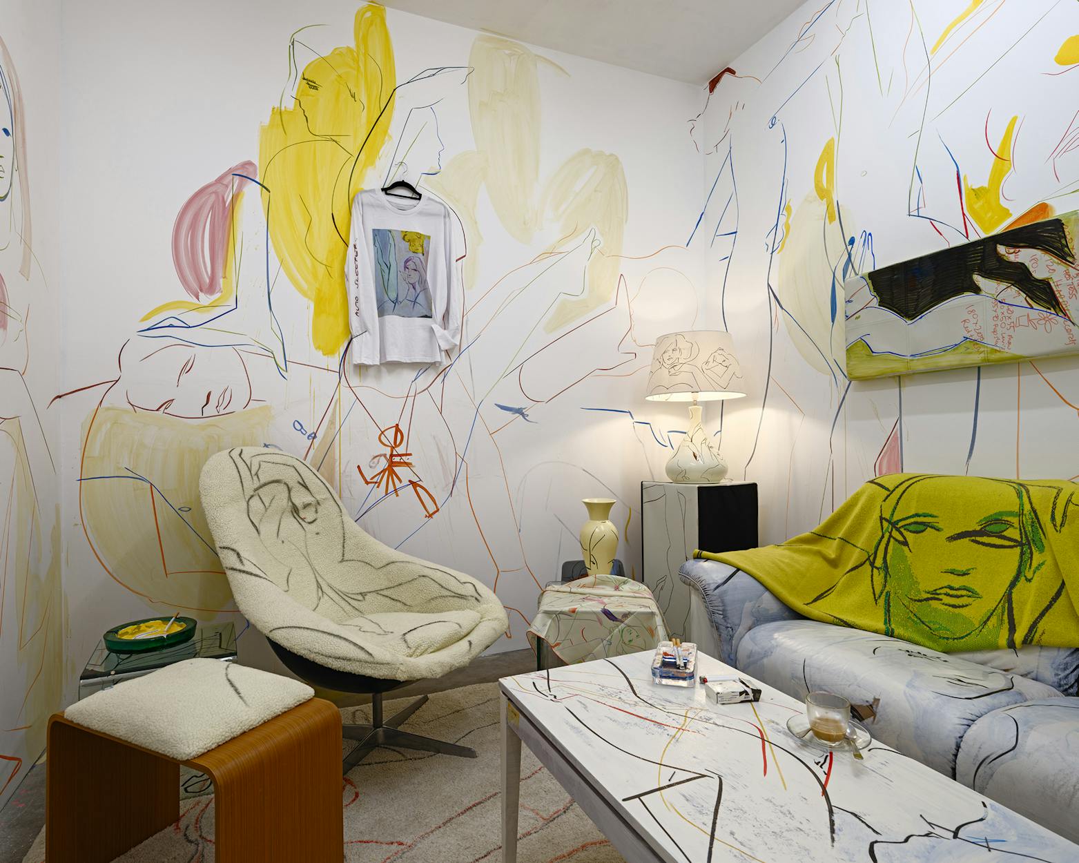 A living room installation with abstract line art of faces and figures painted directly on the walls, a patterned chair, a green throw with a face design on the sofa, a hanging t-shirt, a lamp, small tables, and scattered magazines.