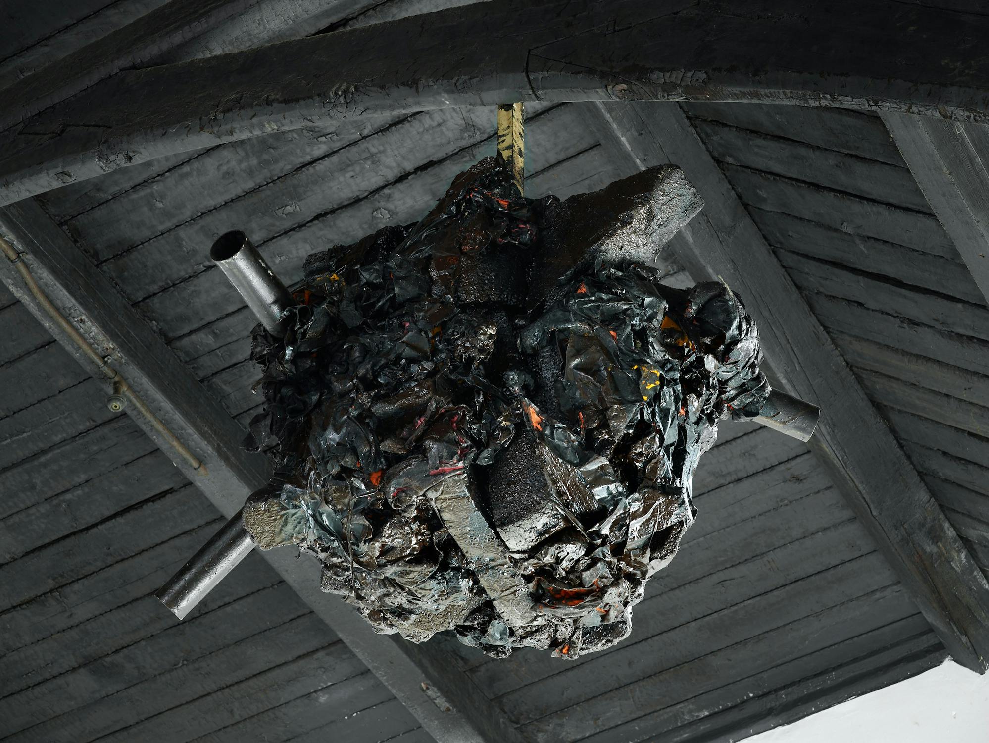 A large, irregular black mass with charred textures and hints of orange and yellow is suspended from a dark, wooden ceiling. Several thick metal pipes jut out from the mass in different directions, creating an industrial, sculptural appearance.