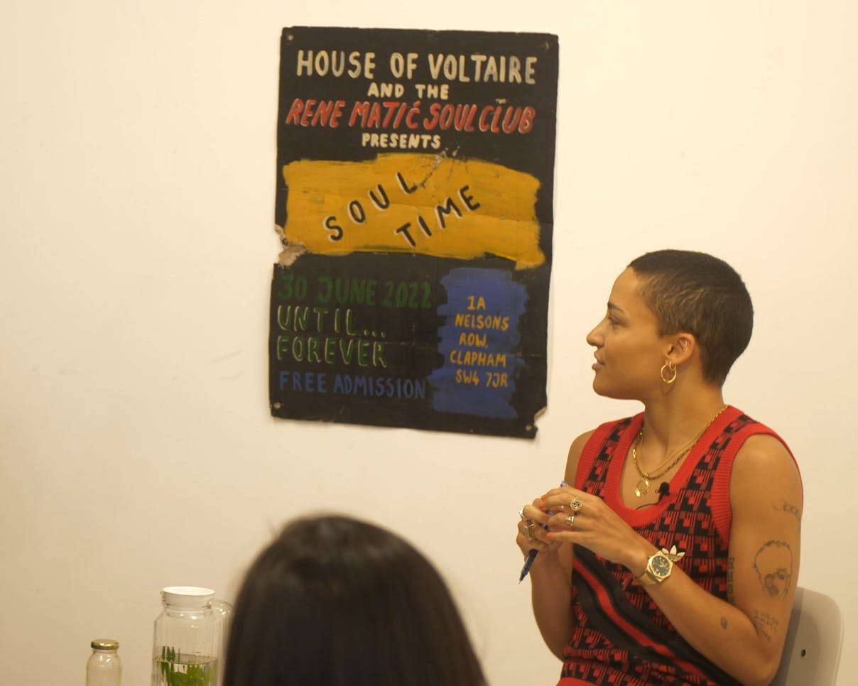 A picture of Rene Matić standing next to their print 'Soul Time' (2022) for House of Voltaire. The artist is looking towards the print over their right shoulder and appears to be speaking to a group standing in the foreground.