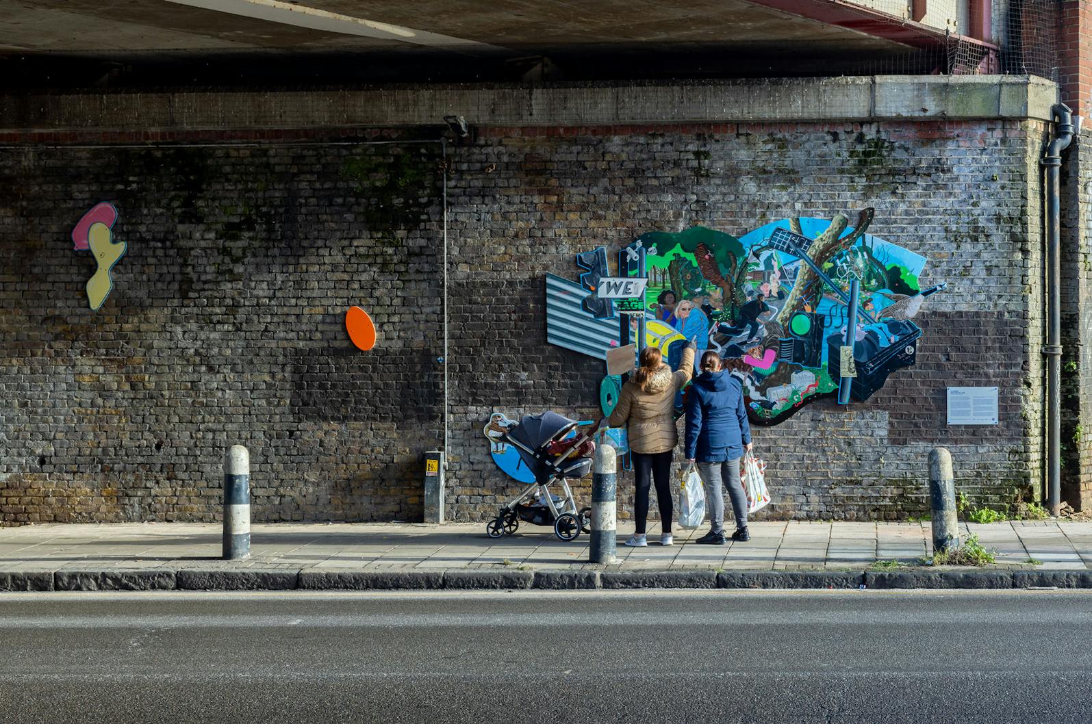 Two women pushing a pushchair stop to look at a vibrant and eclectic mural by Flo Brooks is painted on a brick wall, which features various elements like a man holding a baby, wildlife such as birds, and urban imagery including street signs and graffiti.