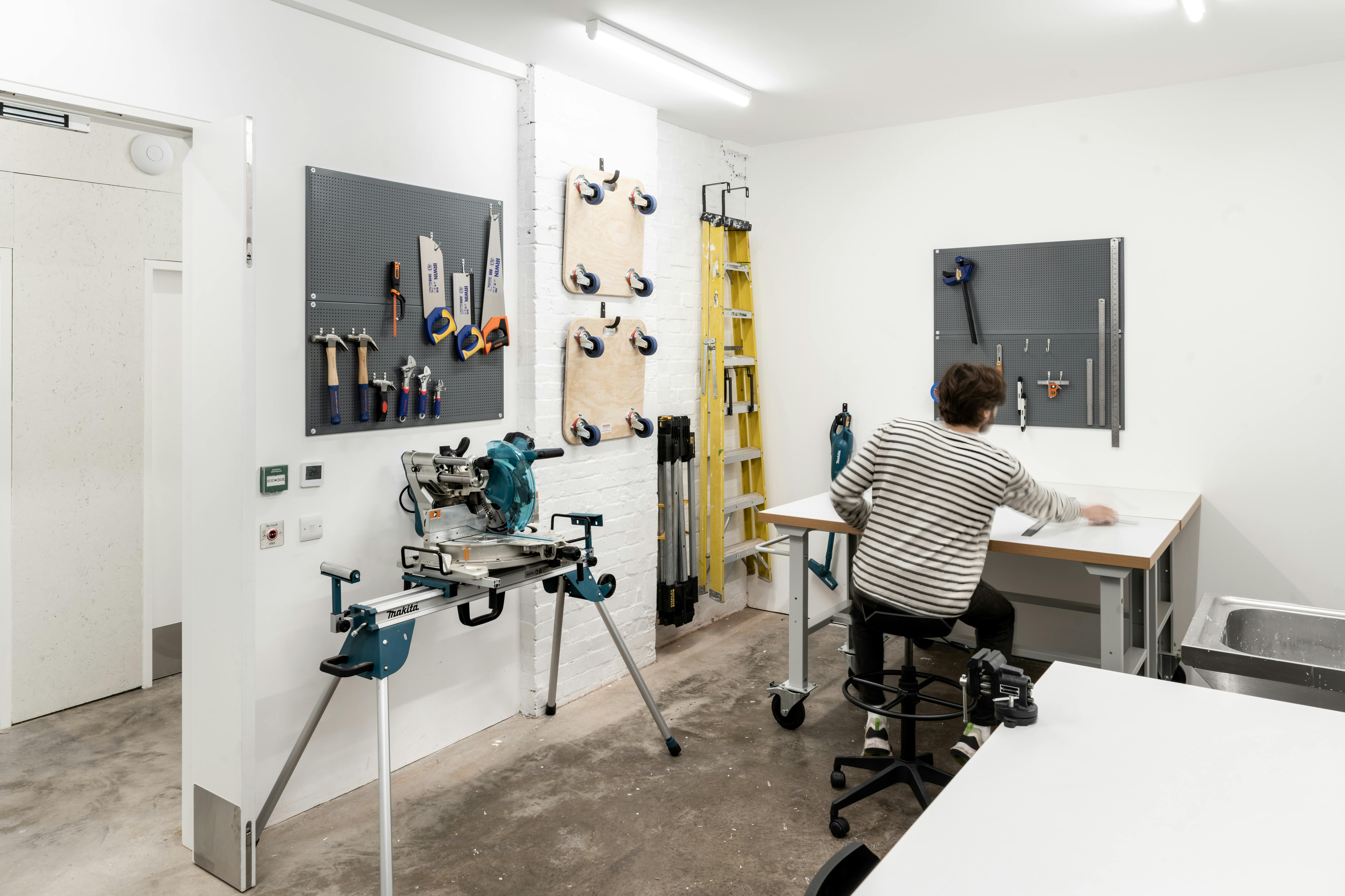 A person with short hair and a striped shirt sits at a workbench in a bright, organized workshop. Tools and clamps hang on pegboards on the walls, a mitre saw is on a stand, and a yellow ladder leans against the back wall. The floor is concrete.