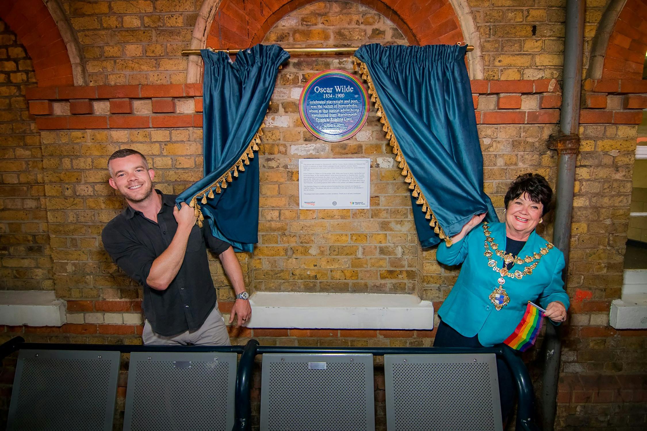 Two people unveil a blue Oscar Wilde plaque on a brick wall. The man on the left smiles, while the woman on the right, wearing a teal jacket and a ceremonial chain, also smiles and holds a rainbow flag.