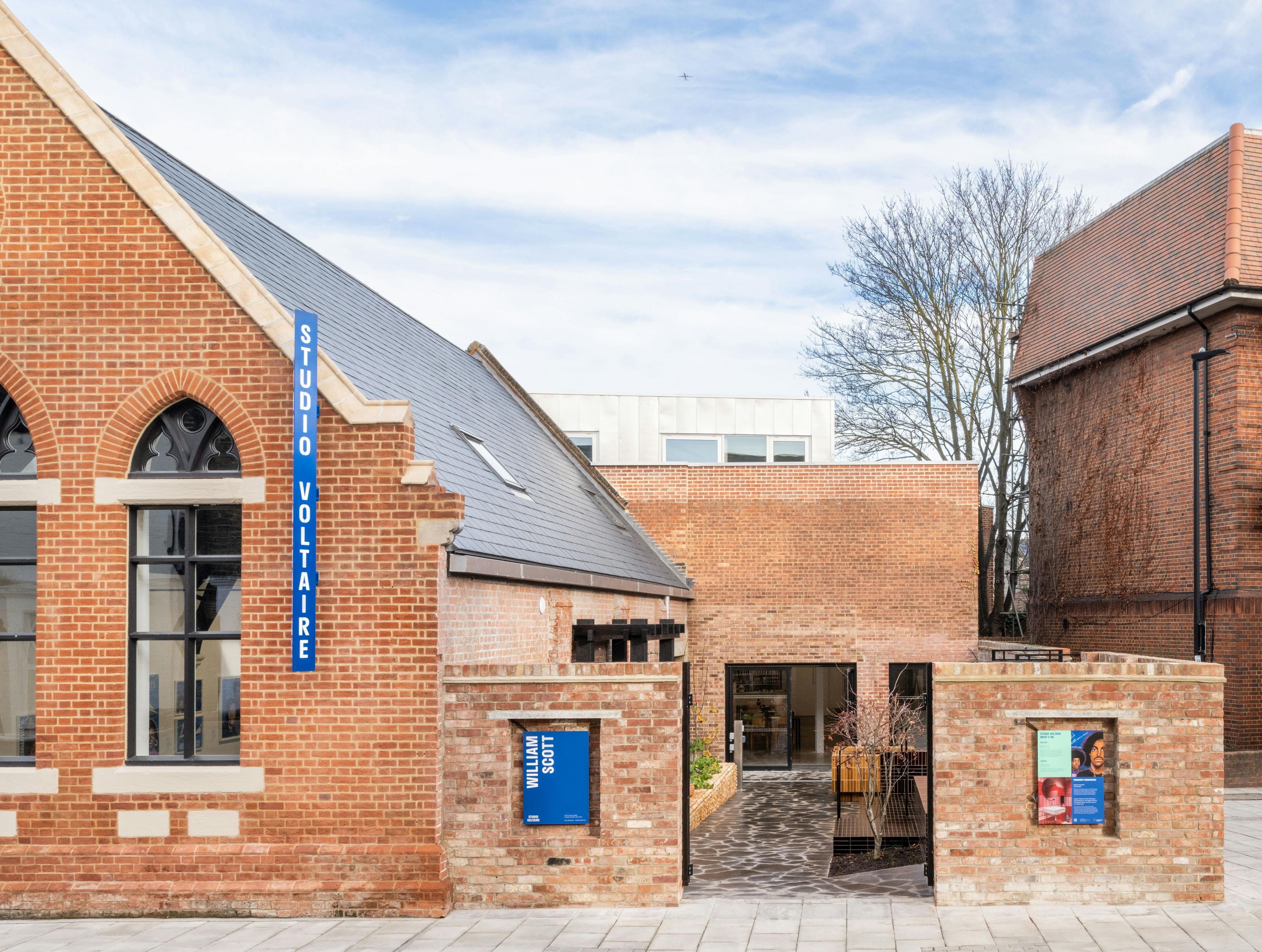 A brick building with a blue "Studio Voltaire" sign, large arched windows and posters by the entrance. The courtyard features modern paving and plants, under a partly cloudy sky.