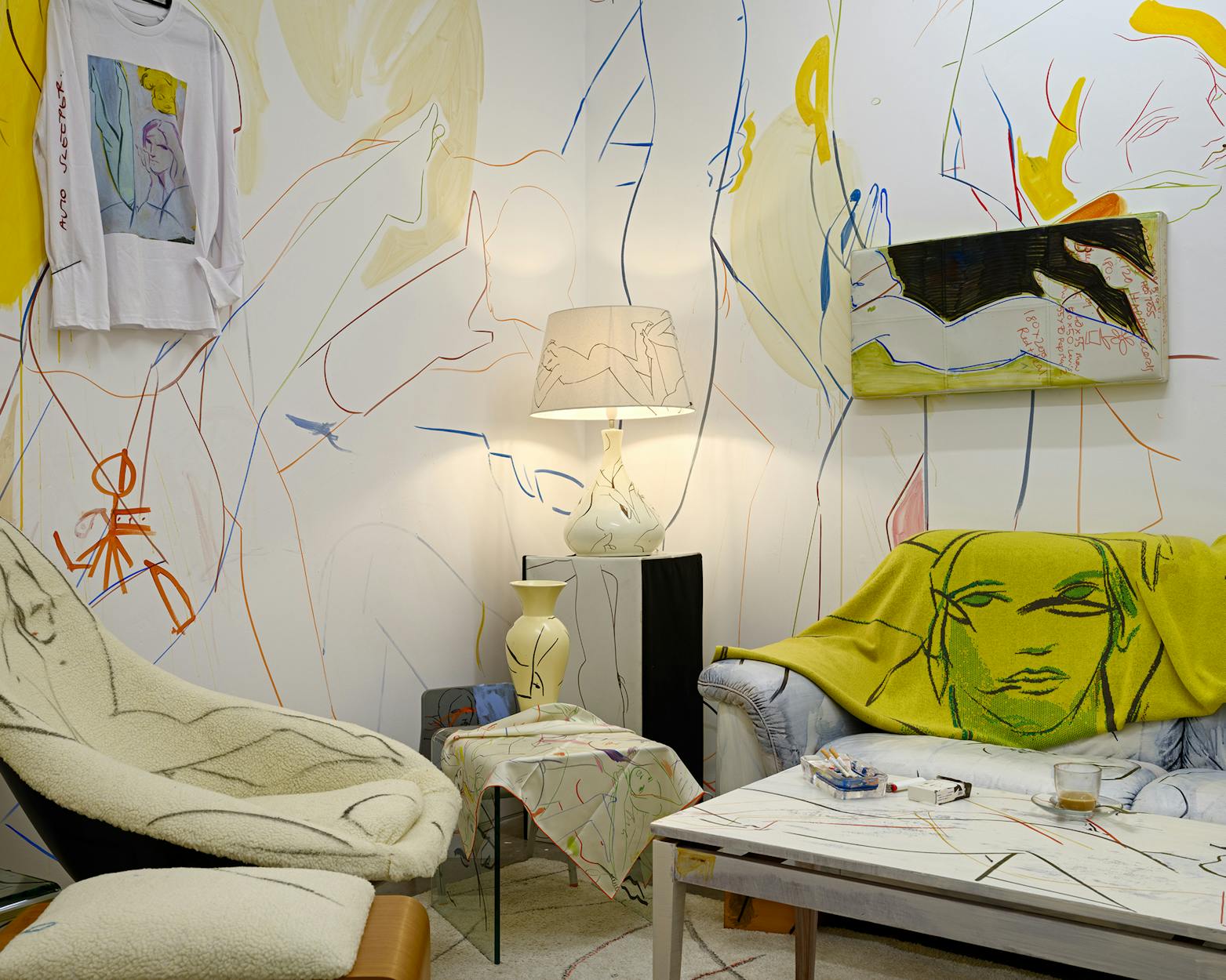 A living room installation with walls, furniture, and decor featuring an abstract line art mural of faces and figures. A yellow throw with a face design covers a sofa, and a matching lamp and t-shirt add to the artistic theme.