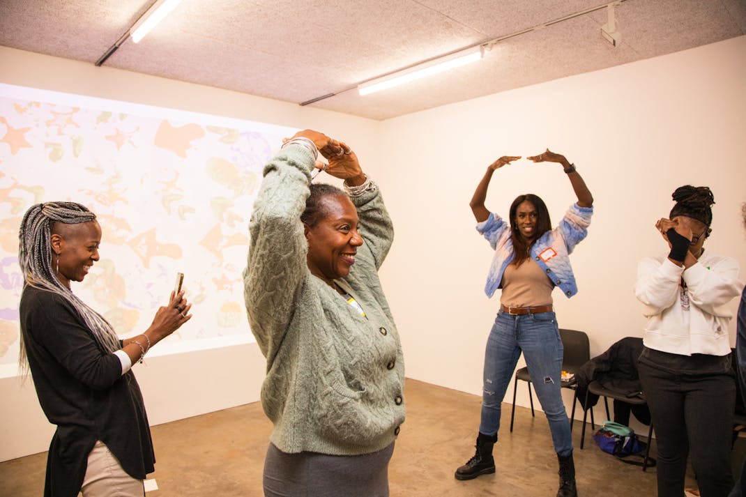 four black women stand in front of a digital projection, posing with their hands above their heads