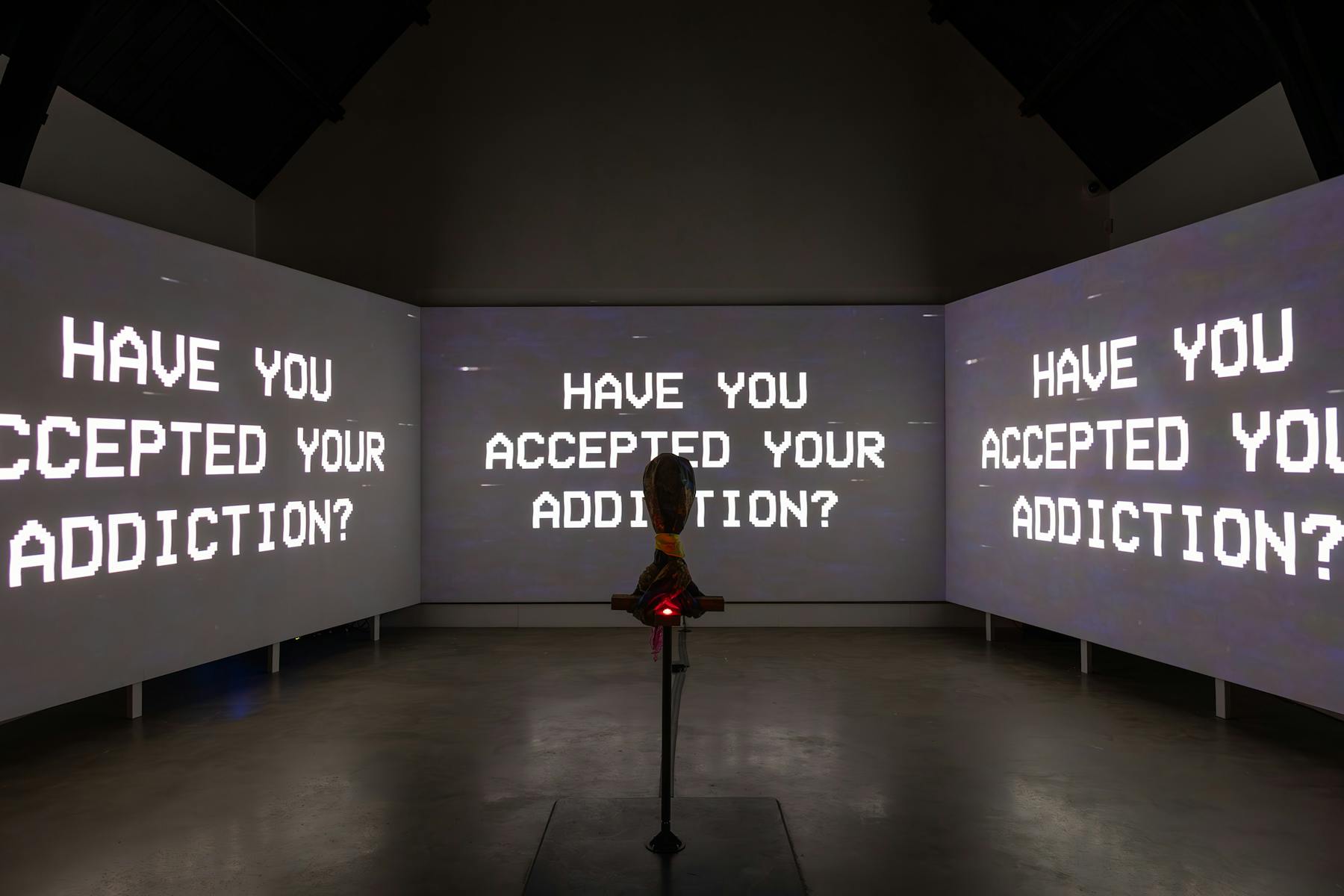 a photograph of three screens that are projected with black and white text. There is text that reads 'HAVE YOU ACCEPTED YOUR ADDICTION'. In the foreground there is a hooded object on a stand. 