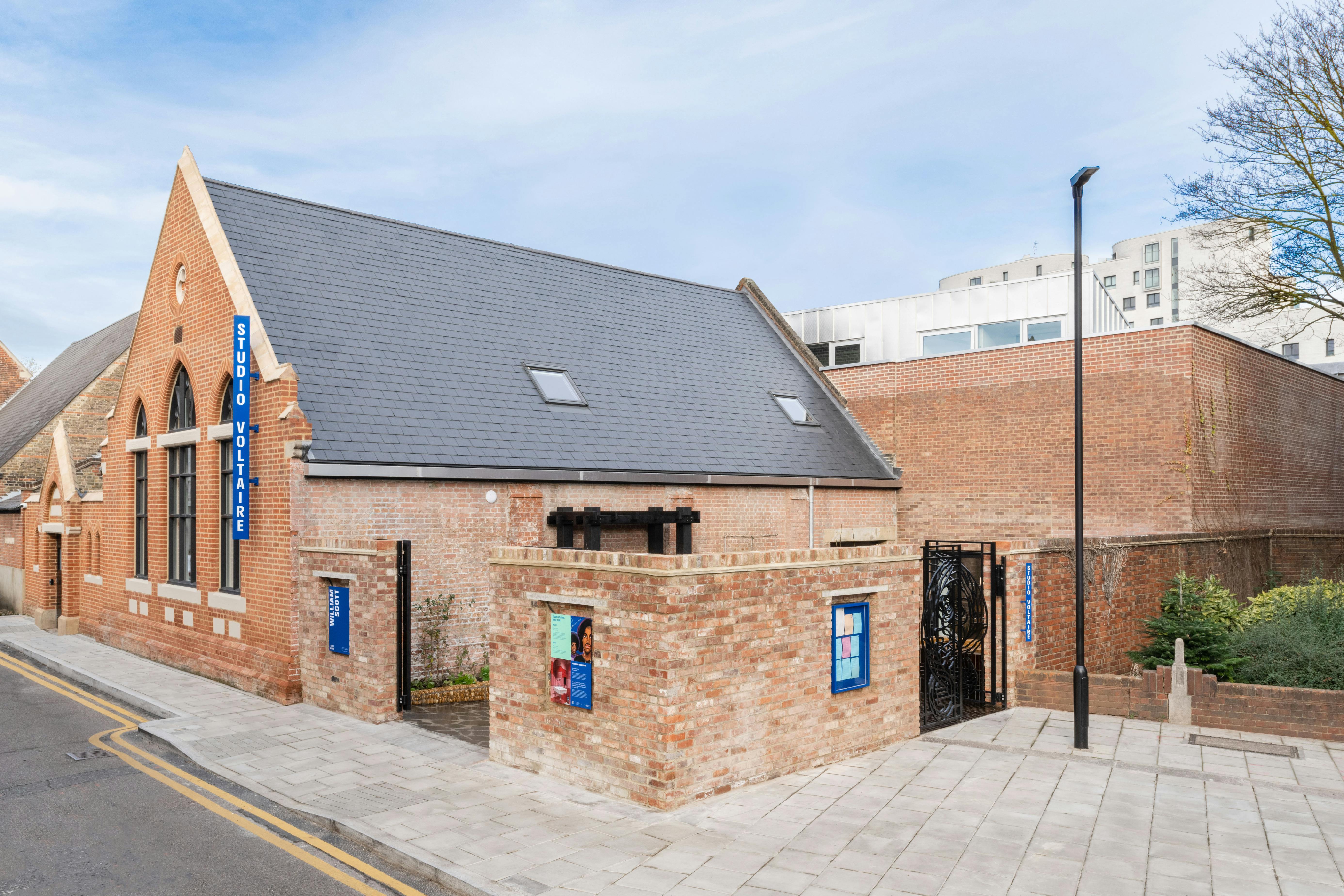 An exterior photograph of Studio Voltaire, taken from the street. The building is a converted Chapel constructed in red brick, with three arched windows and a steep sloped slate roof.